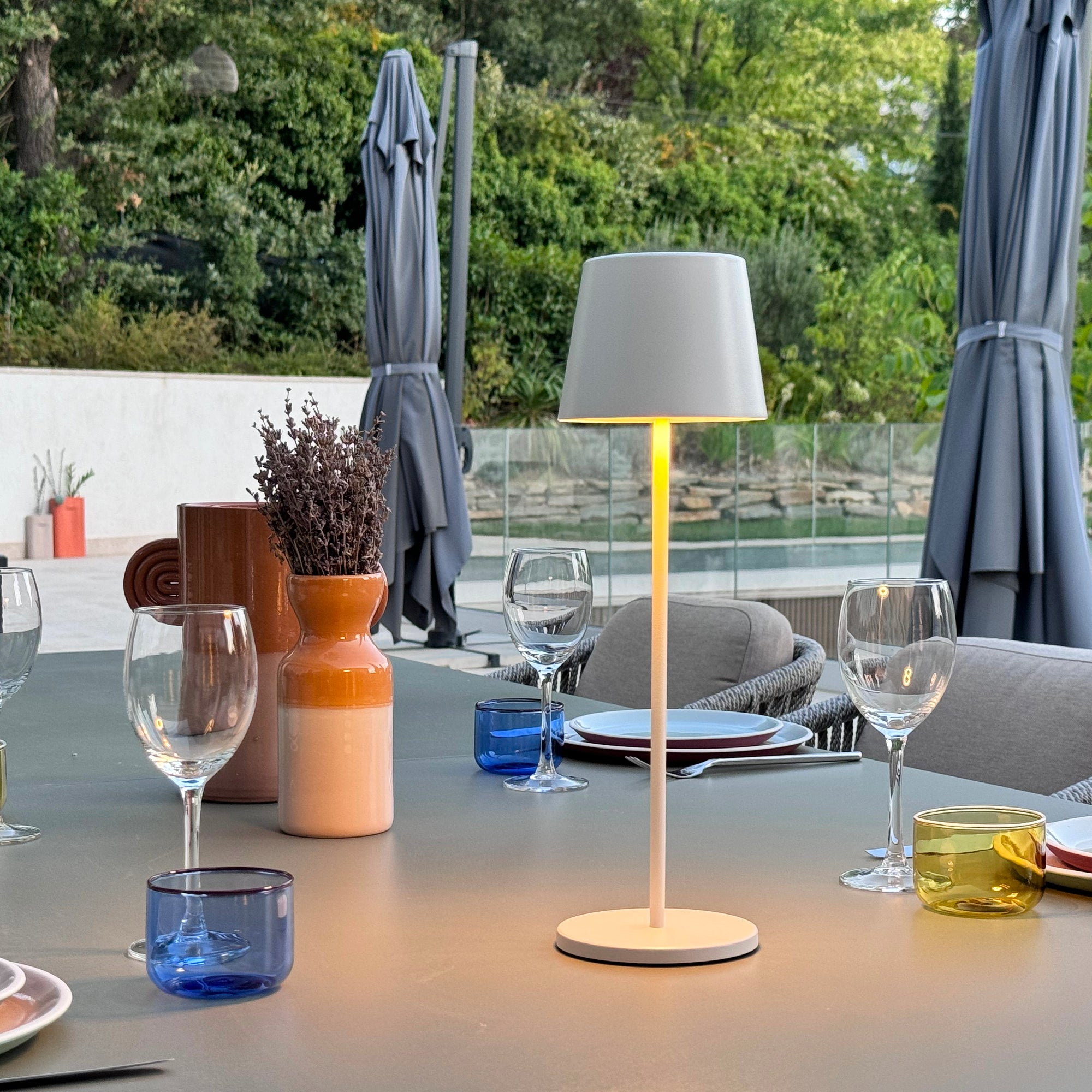 Eine weiße Lampe steht auf einem Tisch mit Gläsern und Geschirr. Der Hintergrund ist ein grüner Garten mit Sonnenschirmen.