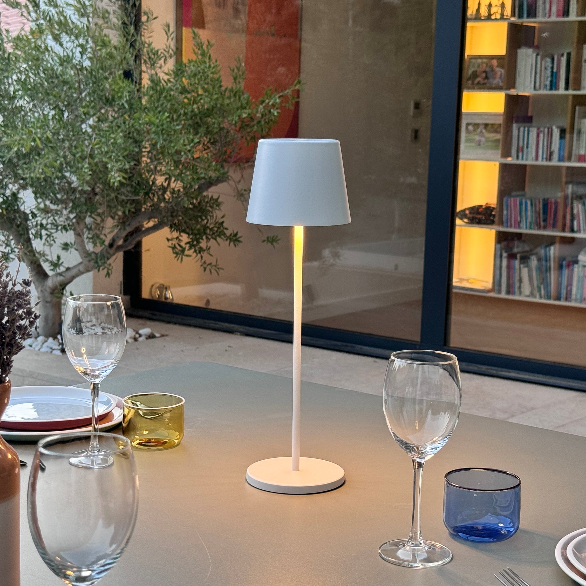 Eine weiße Lampe auf einem Tisch, mit Gläsern und einem Hintergrund einer Szene im Freien.