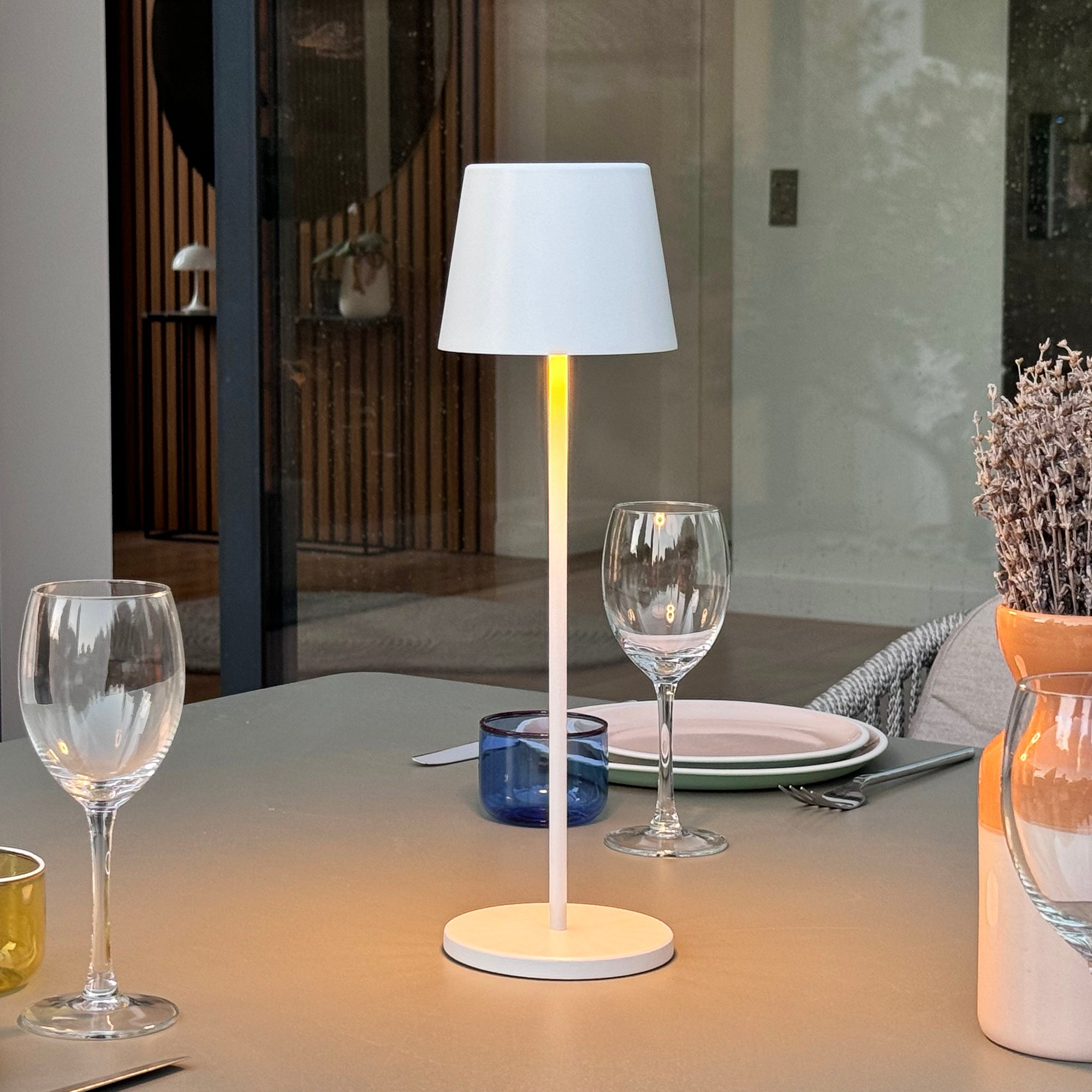 Eine weiße Tischlampe steht auf einem Tisch. Weingläser, ein blaues Glas und Pflanzen stehen auf dem Tisch.