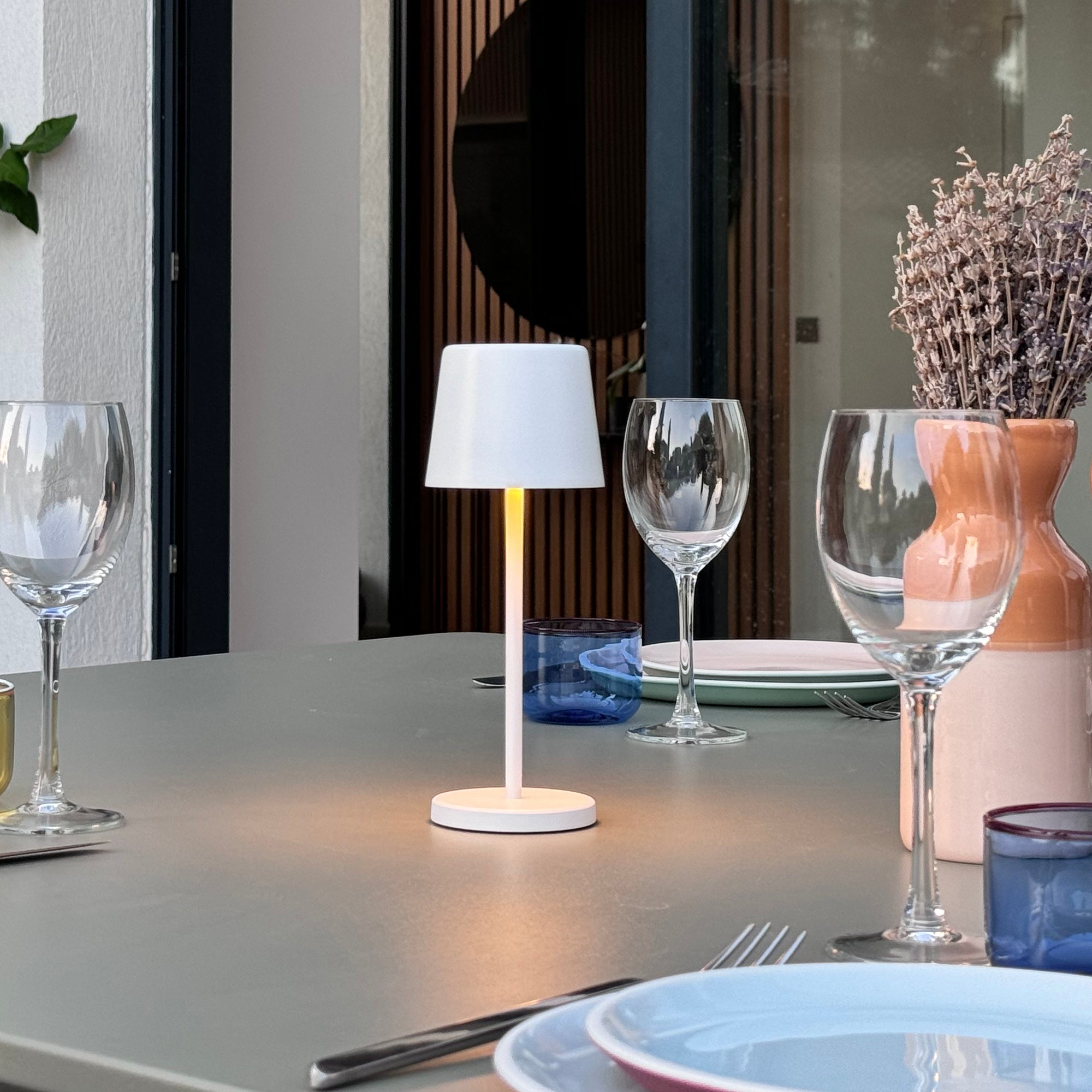 Eine weiße Tischlampe, zwei Weingläser, eine Vase mit Lavendel und blaue Gläser auf einem Tisch.