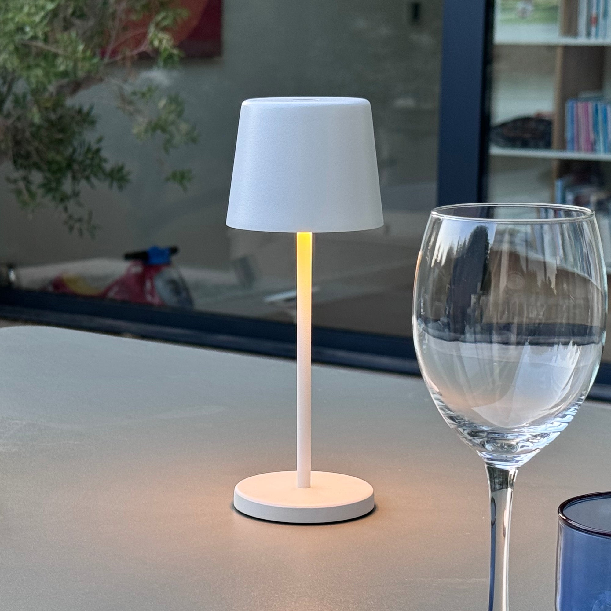 Weiße Lampe auf Tisch, Glas und Außenbereich im Hintergrund.