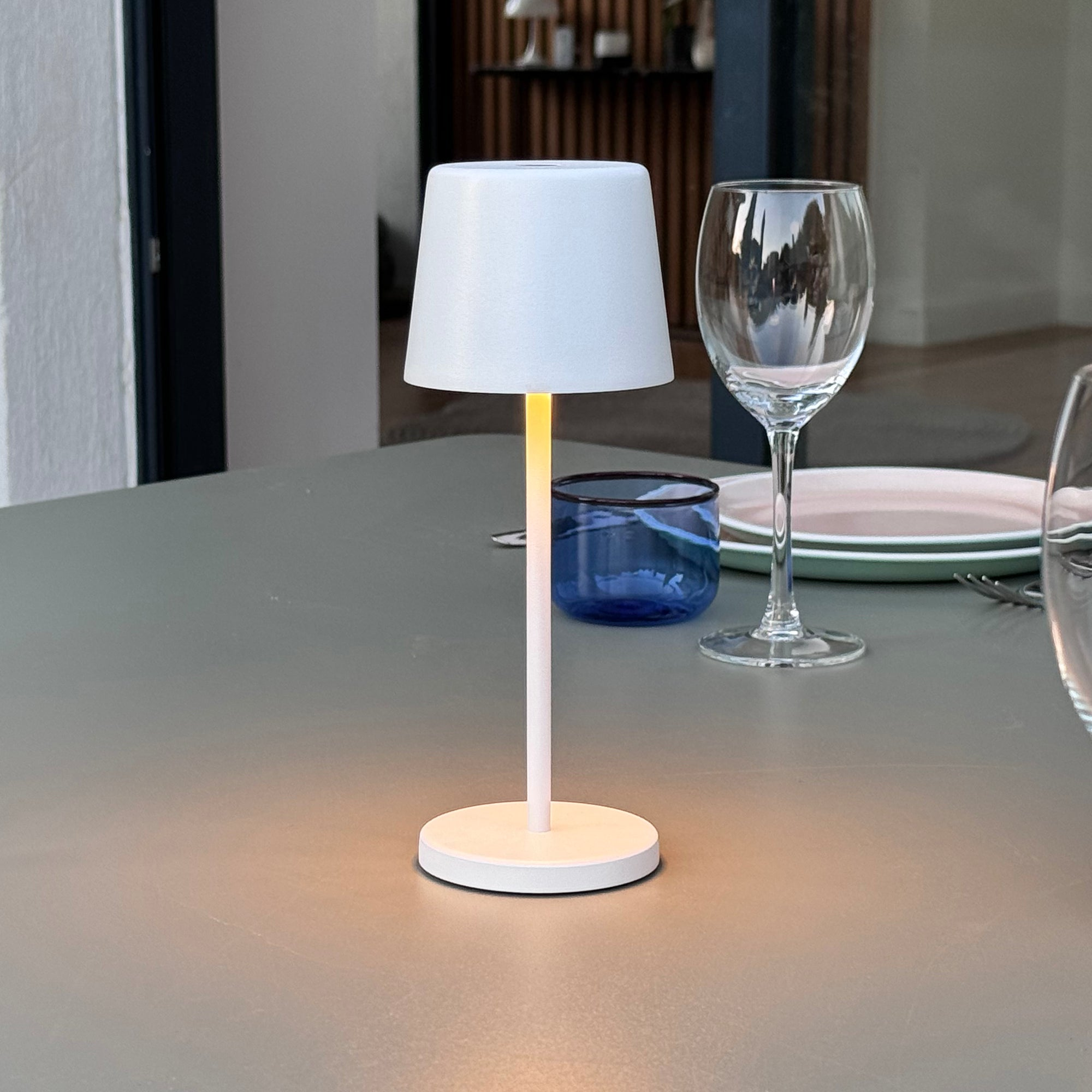 Weiße Tischlampe mit Schirm, steht auf einem Tisch mit Weinglas.