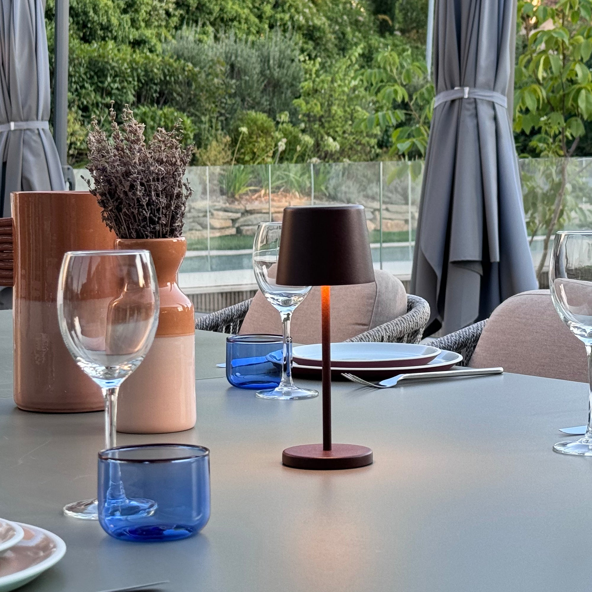 Eine Tischdekoration mit Lampe, Gläsern und Vase in einem Garten mit grünem Laub.