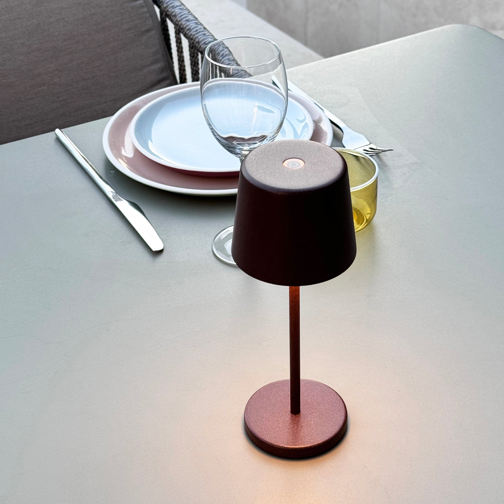 Tisch mit kleiner Lampe, Tellern und Besteck. Die Lampe hat einen dunklen Schirm und einen rötlichen Fuß.