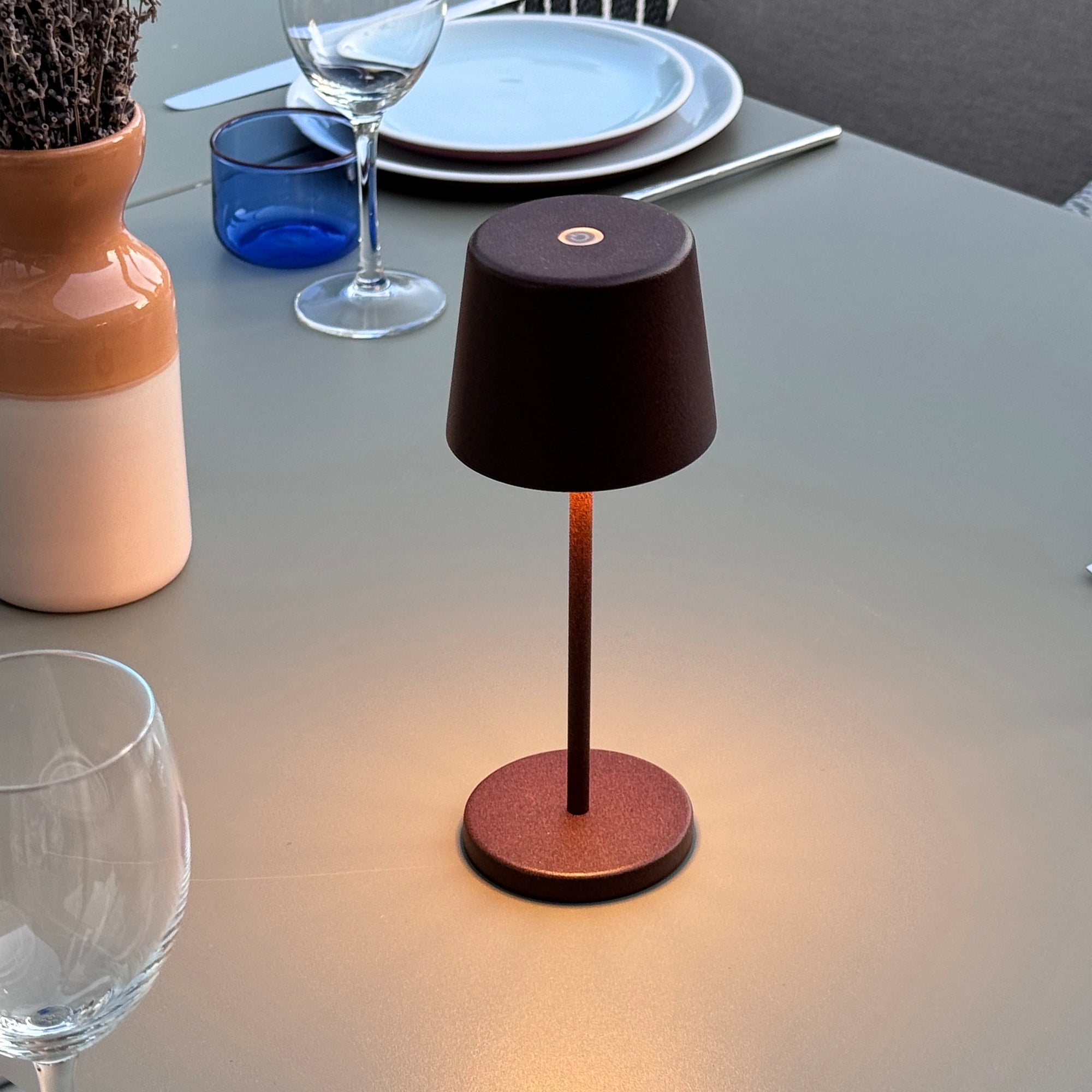 Eine Tischlampe mit einem dunkelbraunen Schirm und Fuß. Die Lampe ist eingeschaltet und wirft einen warmen Schein auf einen Tisch.