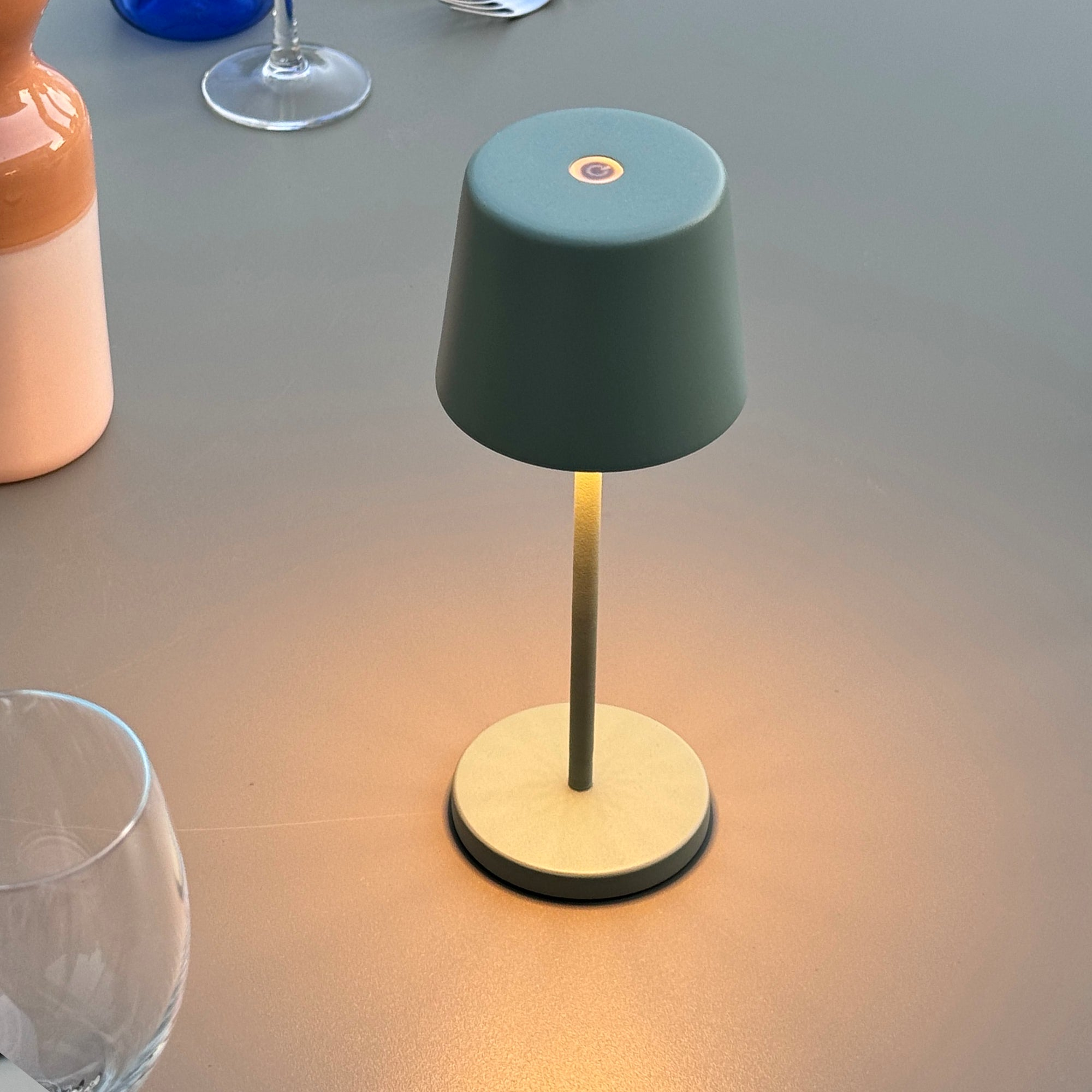 Eine teal Lampe auf einem Tisch. Die Lampe hat einen beigen Sockel und einen teal Schirm. Weiche Beleuchtung.