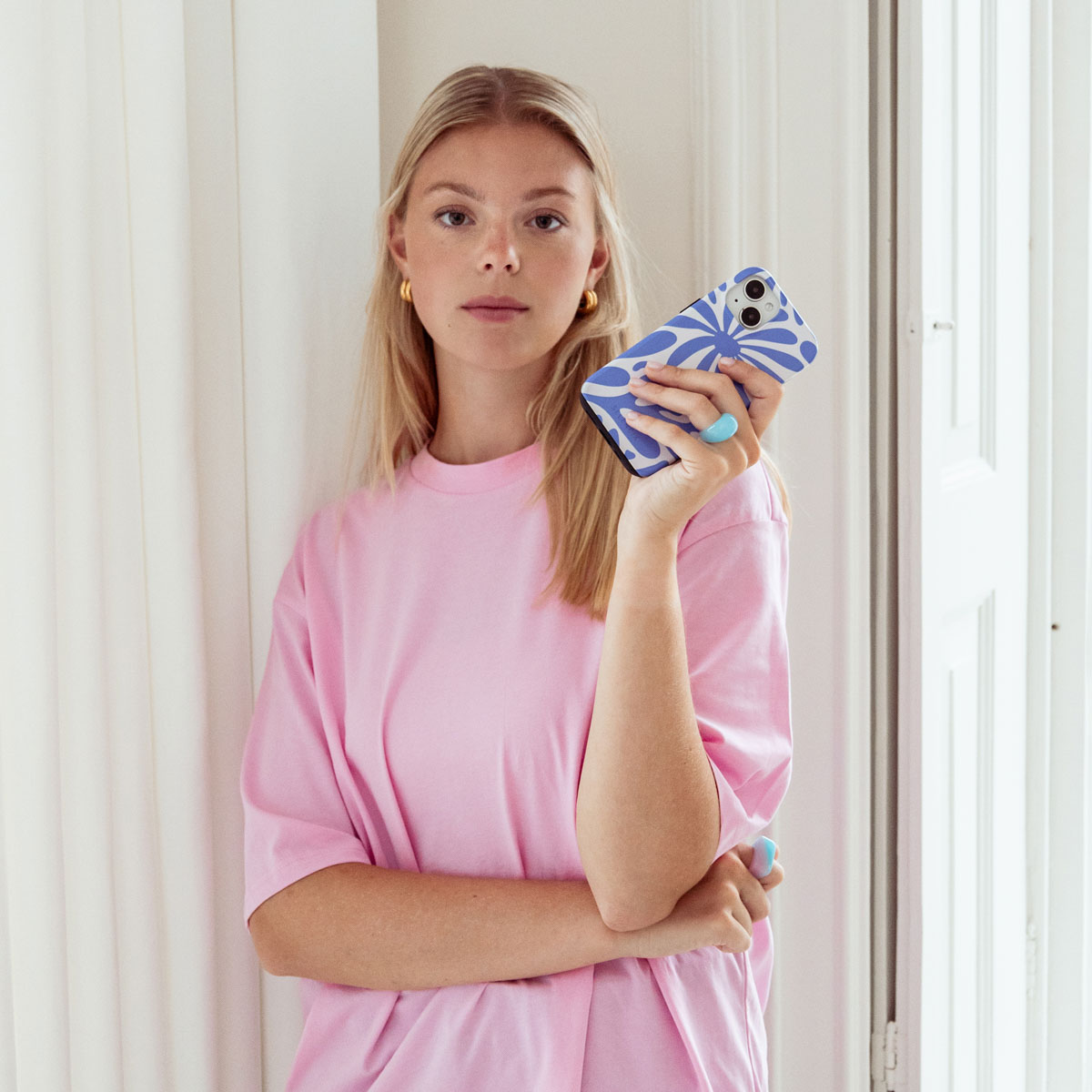 Une femme blonde tient un téléphone dans un étui bleu et blanc, portant une chemise rose, avec des bagues bleues.