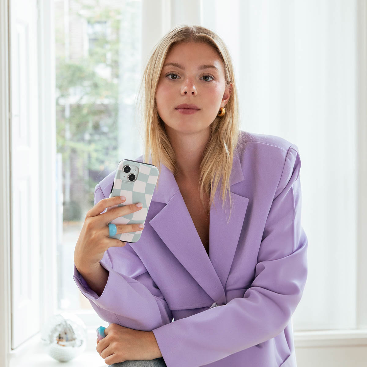Femme blonde tenant un téléphone avec une coque à carreaux verts. Vêtue d'un blazer violet, boucle d'oreille dorée.