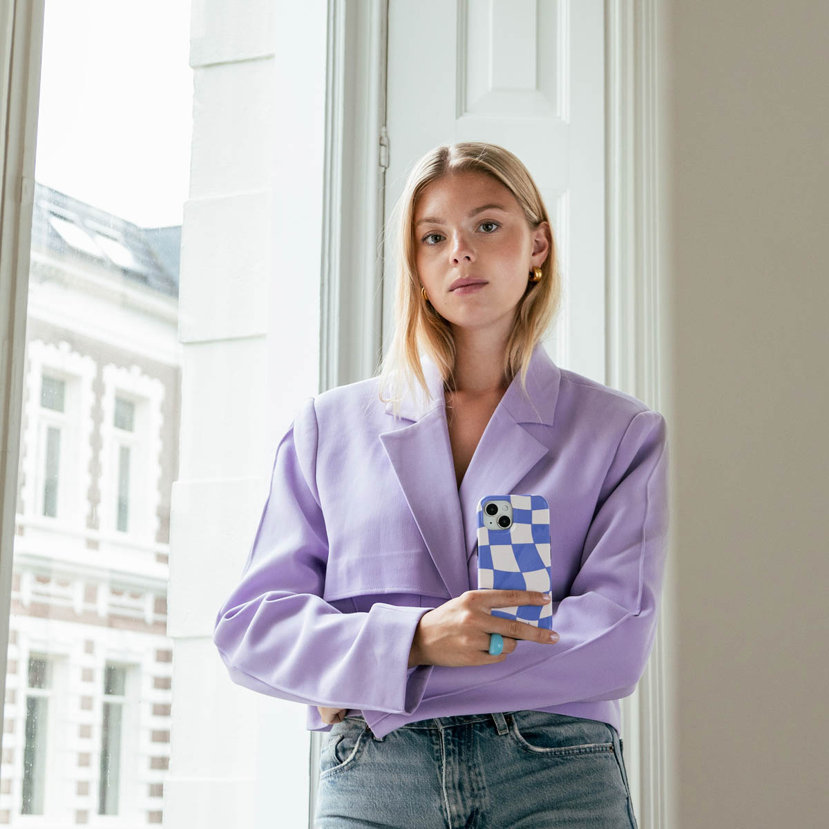 Femme blonde tenant un téléphone avec une coque à carreaux bleus. Vêtue d'un blazer violet.