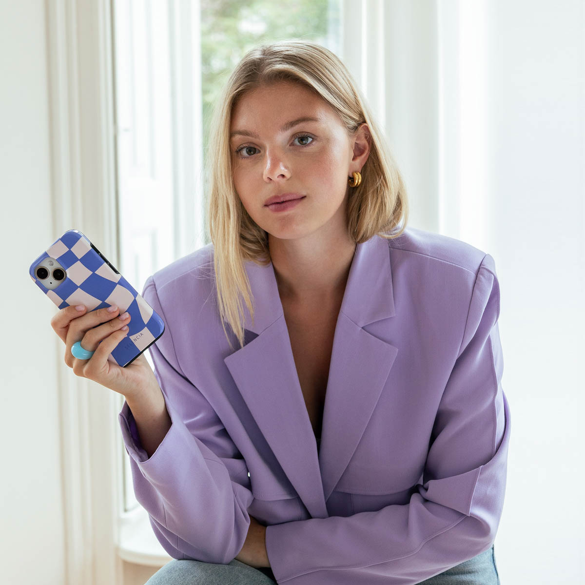 Femme blonde tenant un téléphone avec une coque à carreaux bleus. Vêtue d'un blazer violet, boucles d'oreilles dorées.