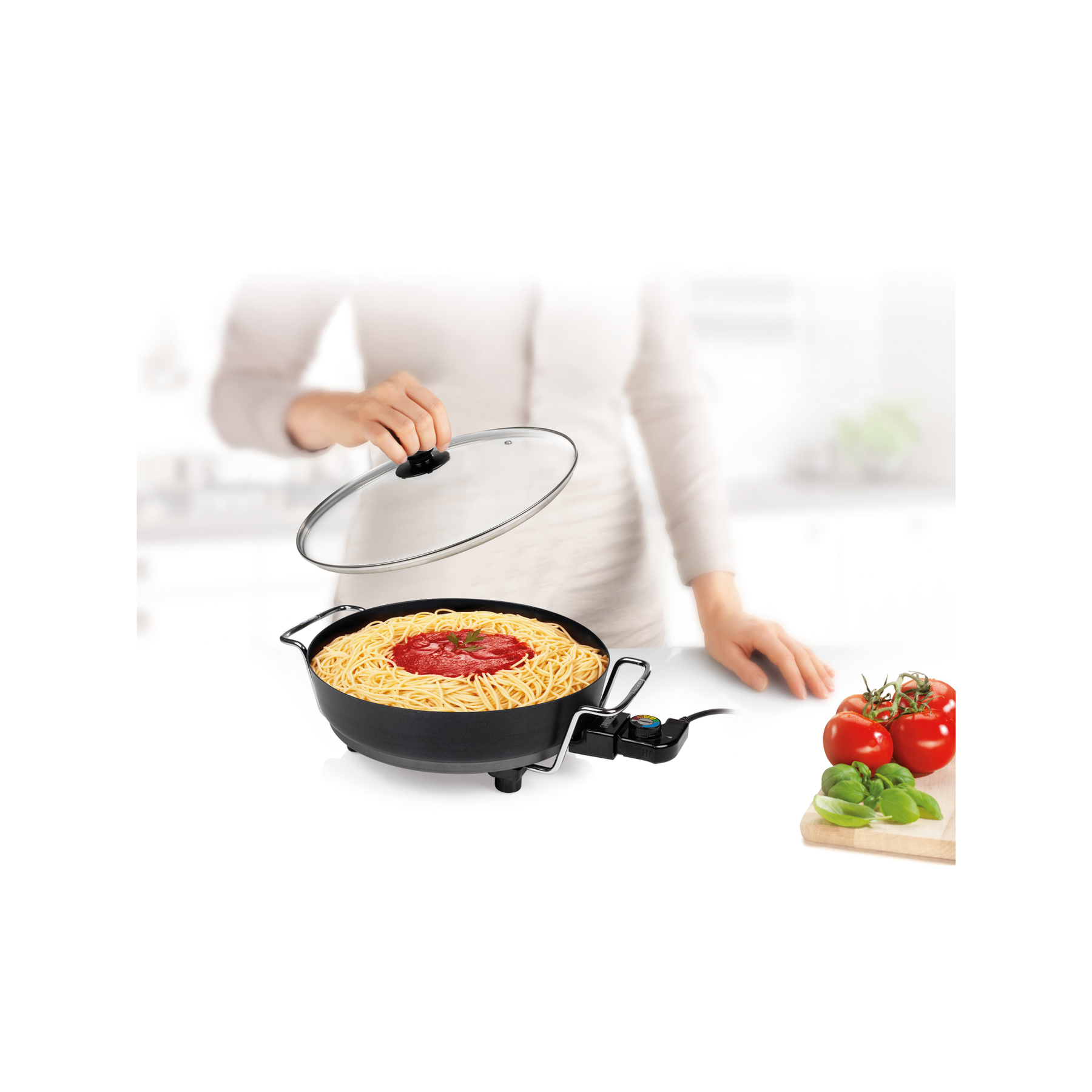 Persoon met pasta in een pan, tomaten en basilicumblaadjes op een wit oppervlak.