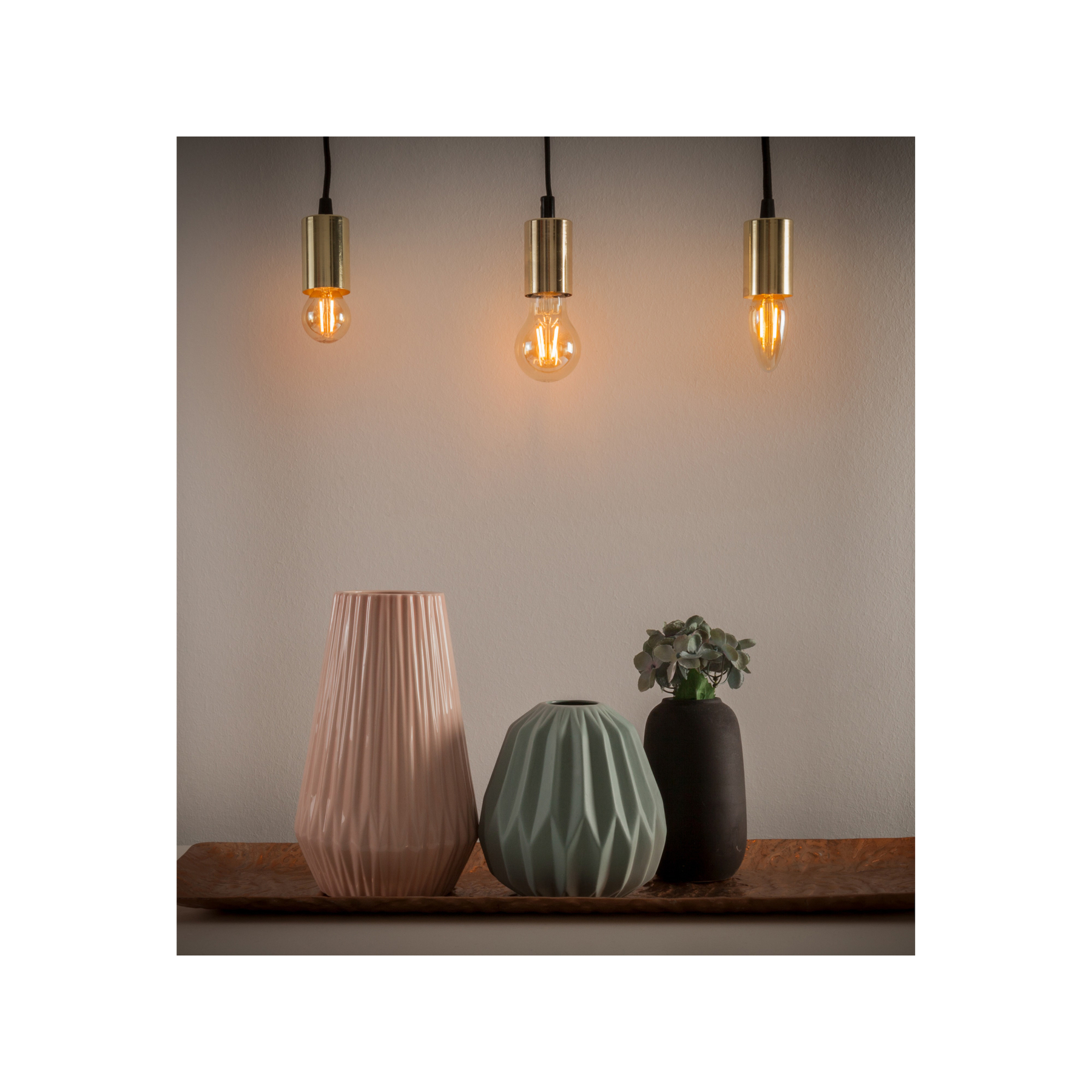 Trzy wiszące żarówki nad wazonami. Różowe, zielone i czarne wazony na tacy.