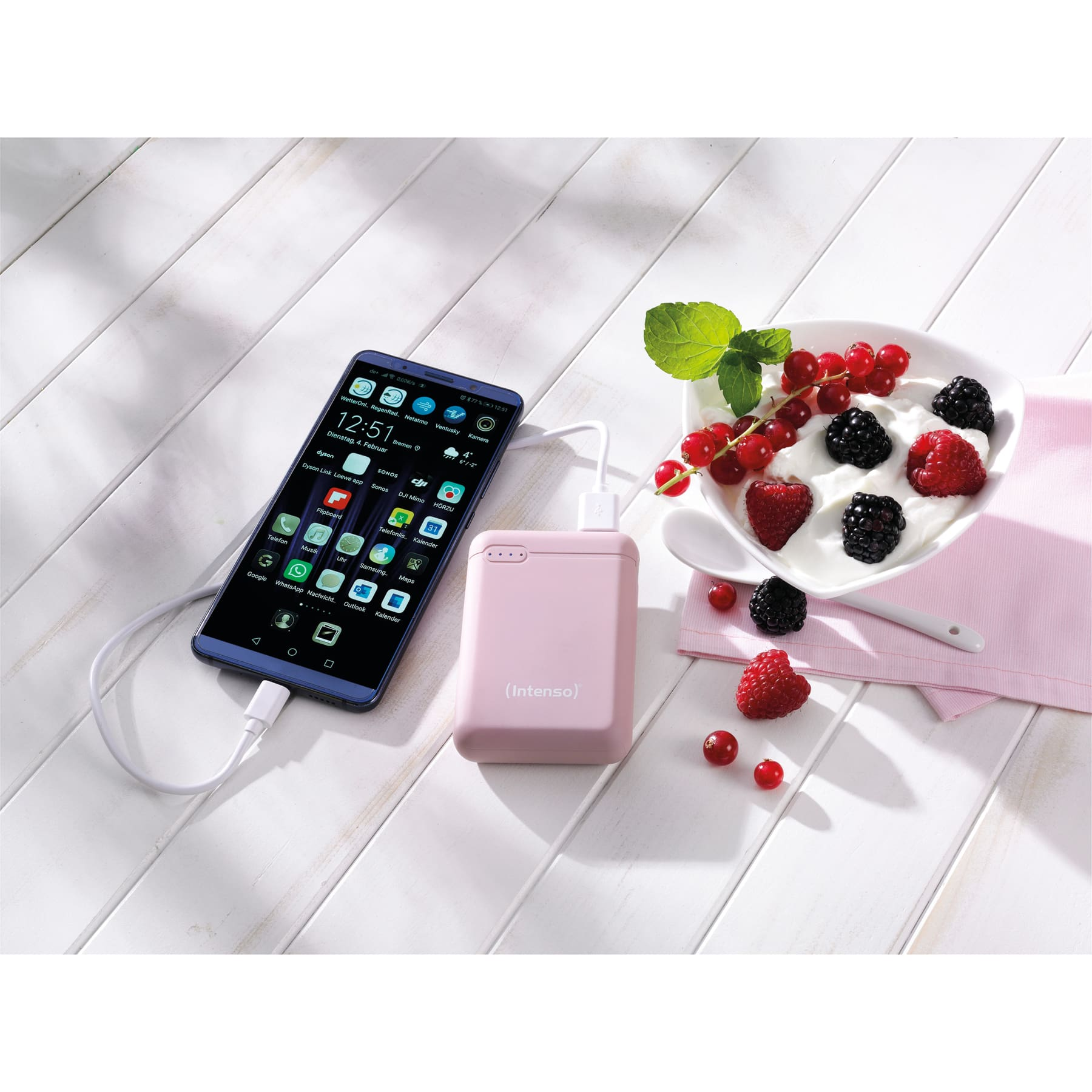 Ein Mobiltelefon, das neben einer Powerbank geladen wird. Rechts ist eine Schale mit Beeren.