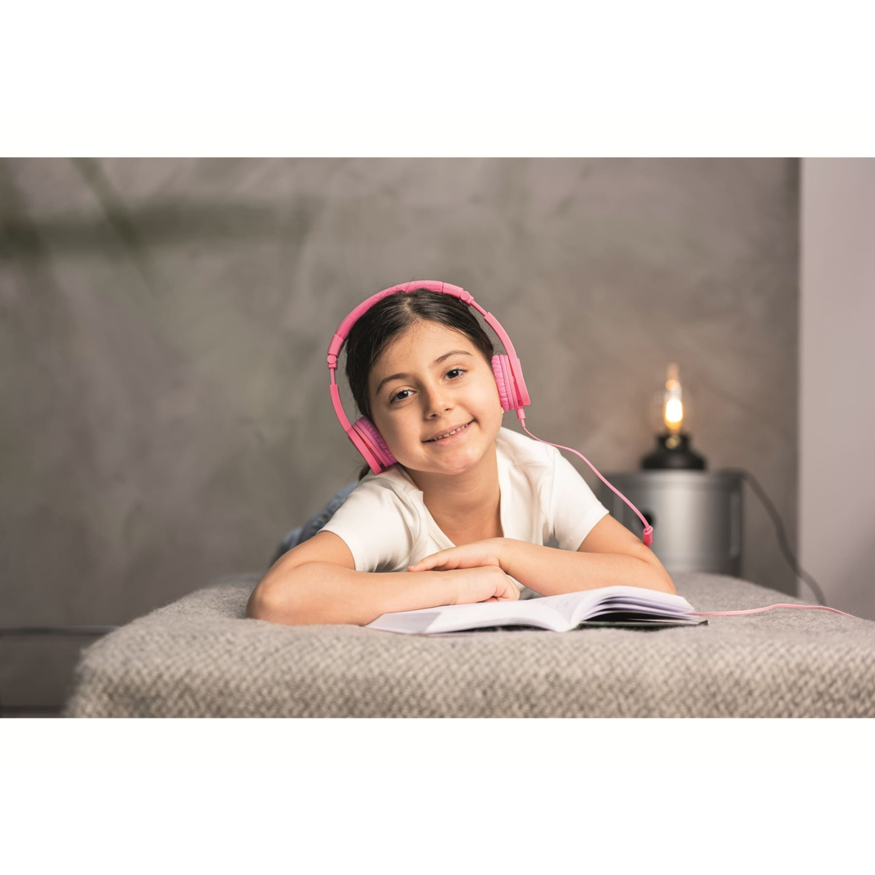 Ragazza sul letto con cuffie rosa, legge un libro. Lampada di lato.
