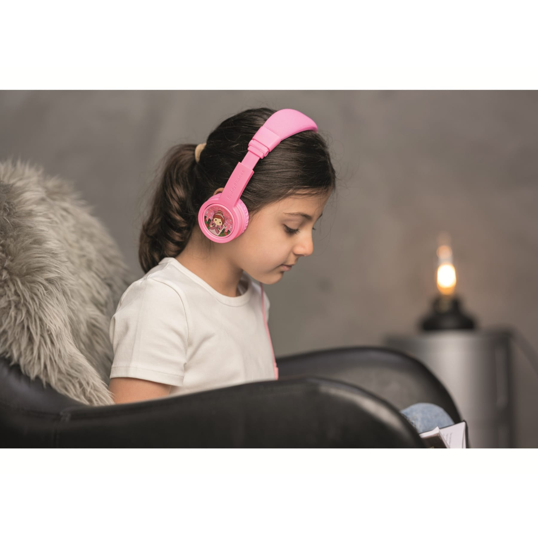Ragazza con cuffie rosa, legge un libro. Sfondo: sedia scura, coperta soffice e lampada accesa.