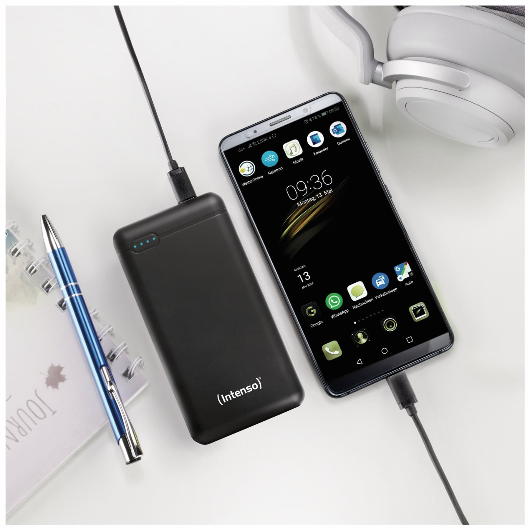 Smartphone, der von einer schwarzen Powerbank aufgeladen wird, mit Stift und Notizbuch auf einem weißen Tisch.