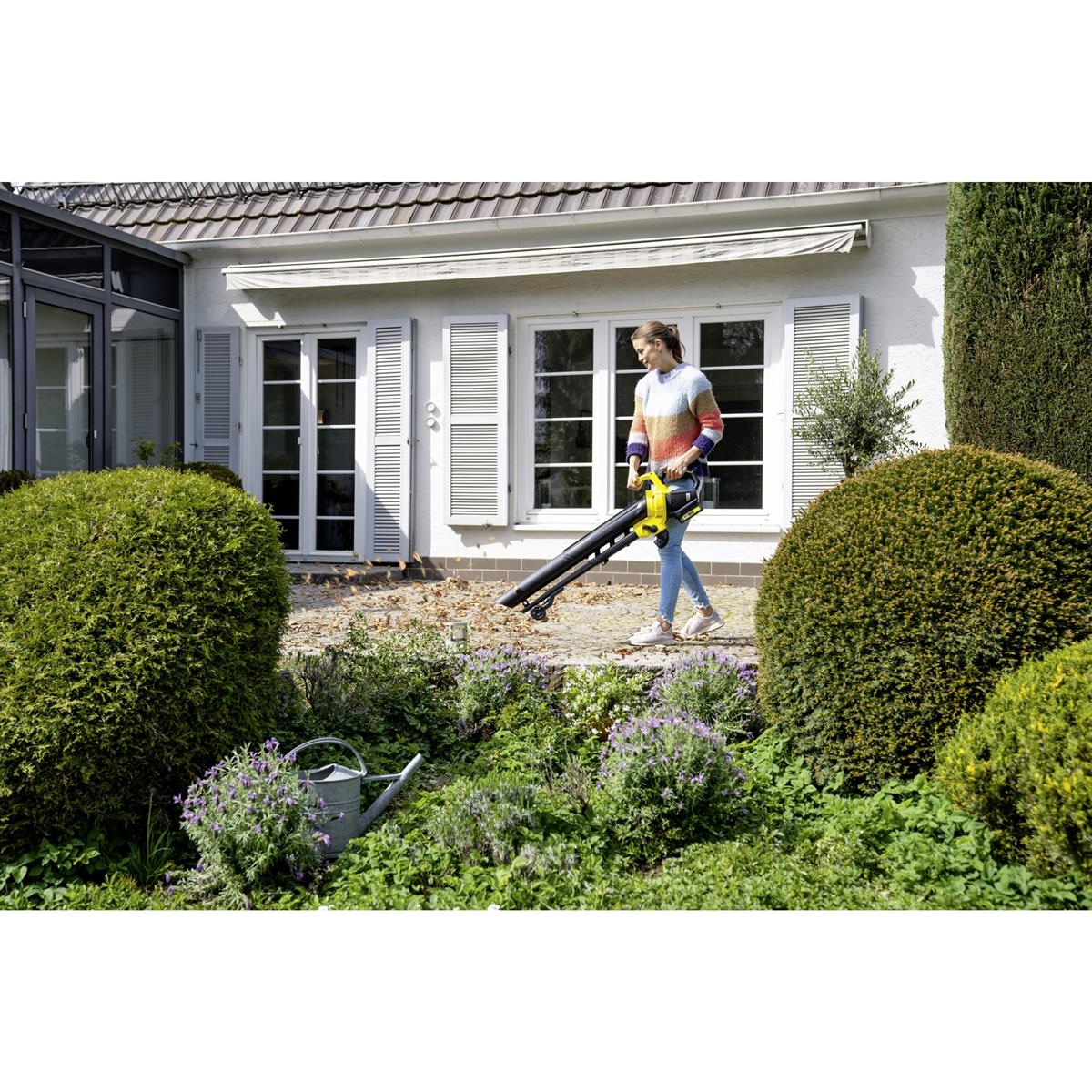 Kobieta używająca dmuchawy do liści w ogrodzie, z domem w tle.