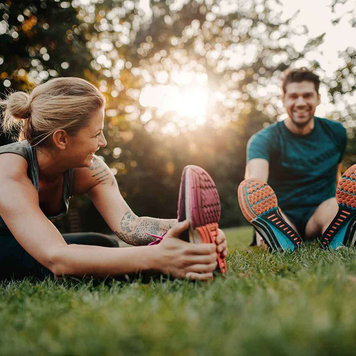 Due persone che si allungano le gambe sull'erba. Una donna ha tatuaggi. Sfondo verde.