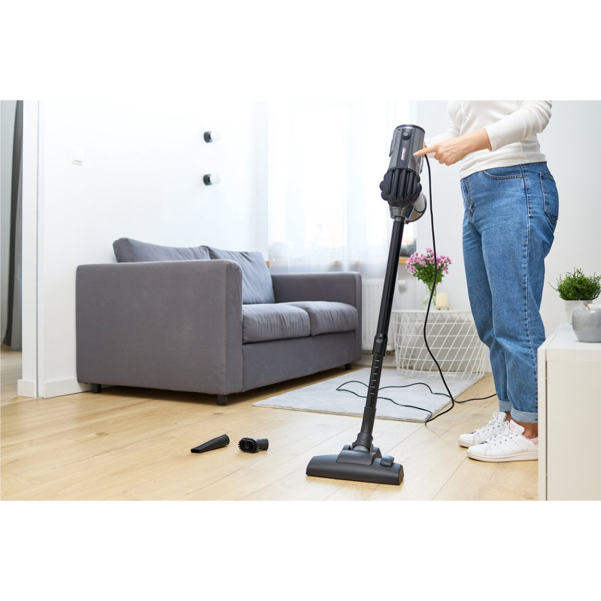 Eine Frau in Jeans saugt einen Raum mit einem schwarzen Staubsauger. Vordergrund: Staubsaugerteile. Hintergrund: Sofa.