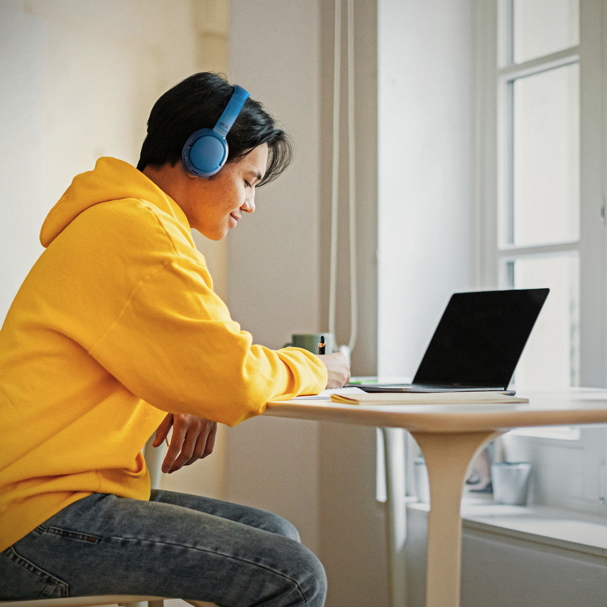 Eine Person mit Kopfhörern und gelbem Kapuzenpullover schreibt an einem Schreibtisch mit Laptop.