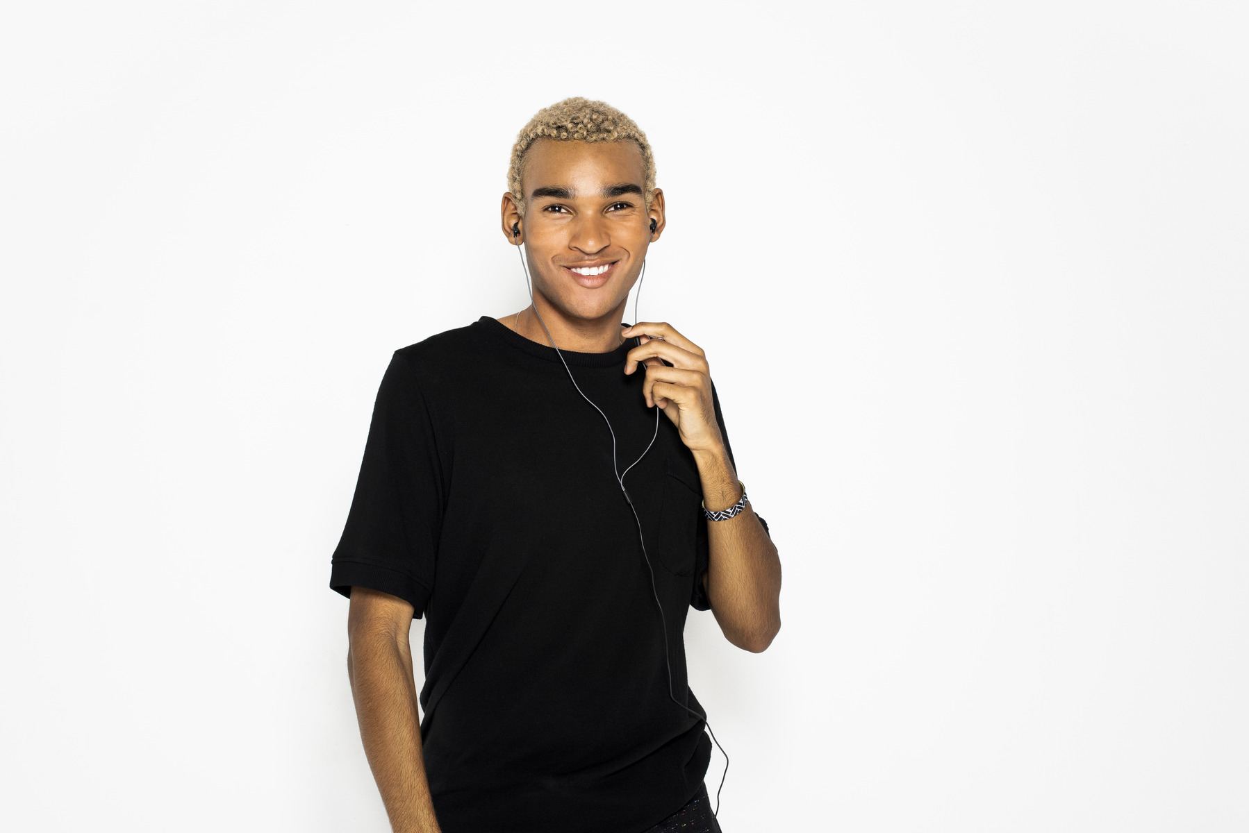 Ein lächelnder Mann mit blonden Haaren, der ein schwarzes T-Shirt trägt und vor weißem Hintergrund kabelgebundene Ohrhörer hält.