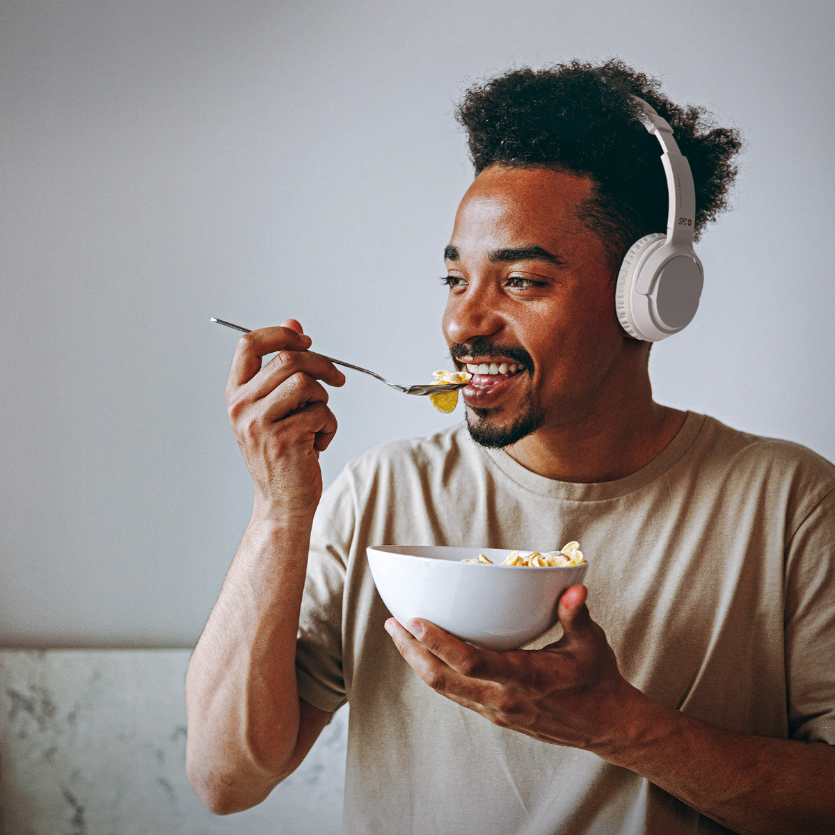 Ein Mann mit Kopfhörern isst Müsli aus einer Schüssel. Er lächelt und hält einen Löffel. Der Hintergrund ist hellgrau.