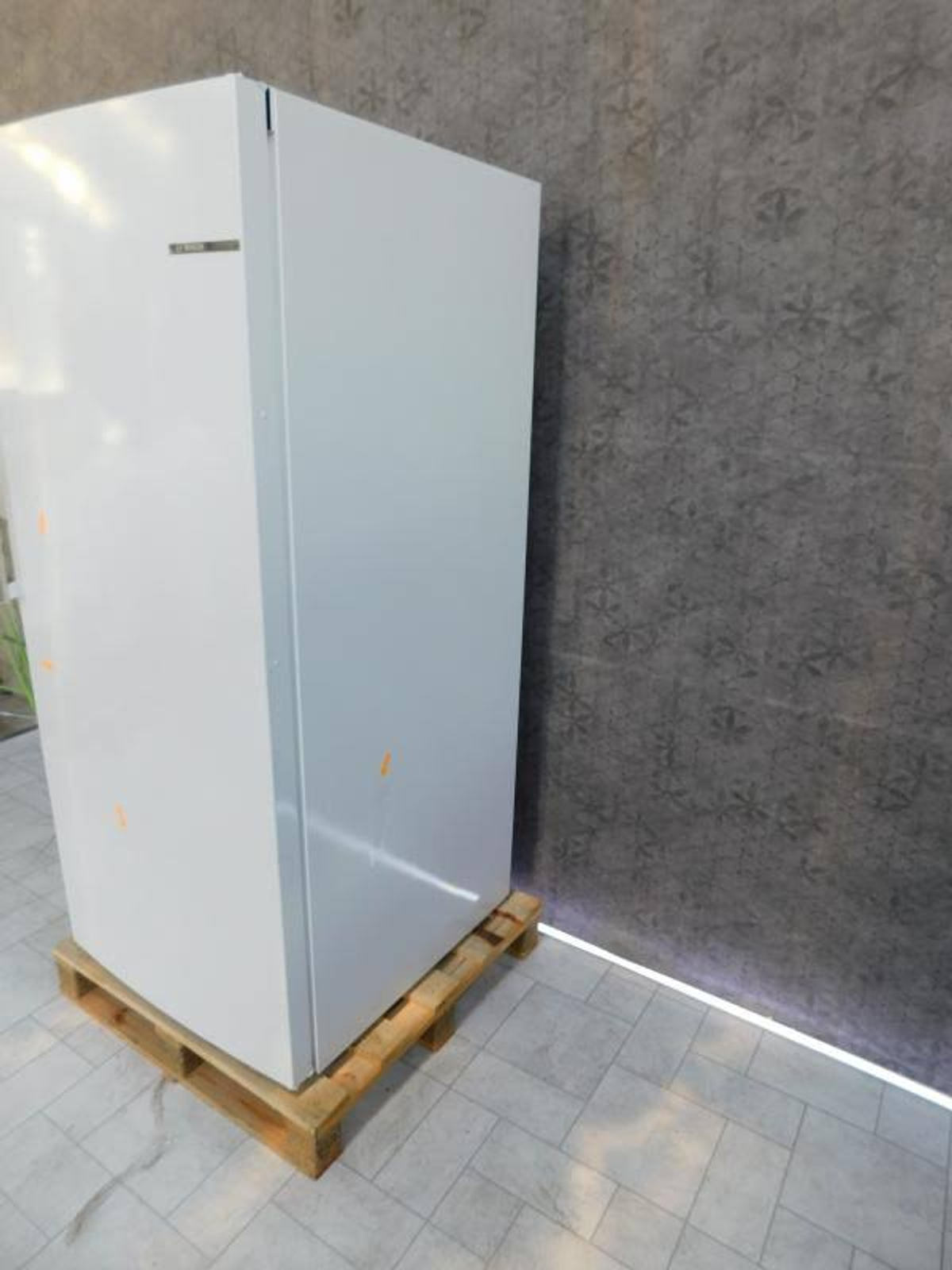 Biała lodówka na drewnianej palecie na tle teksturowanej szarej ściany.