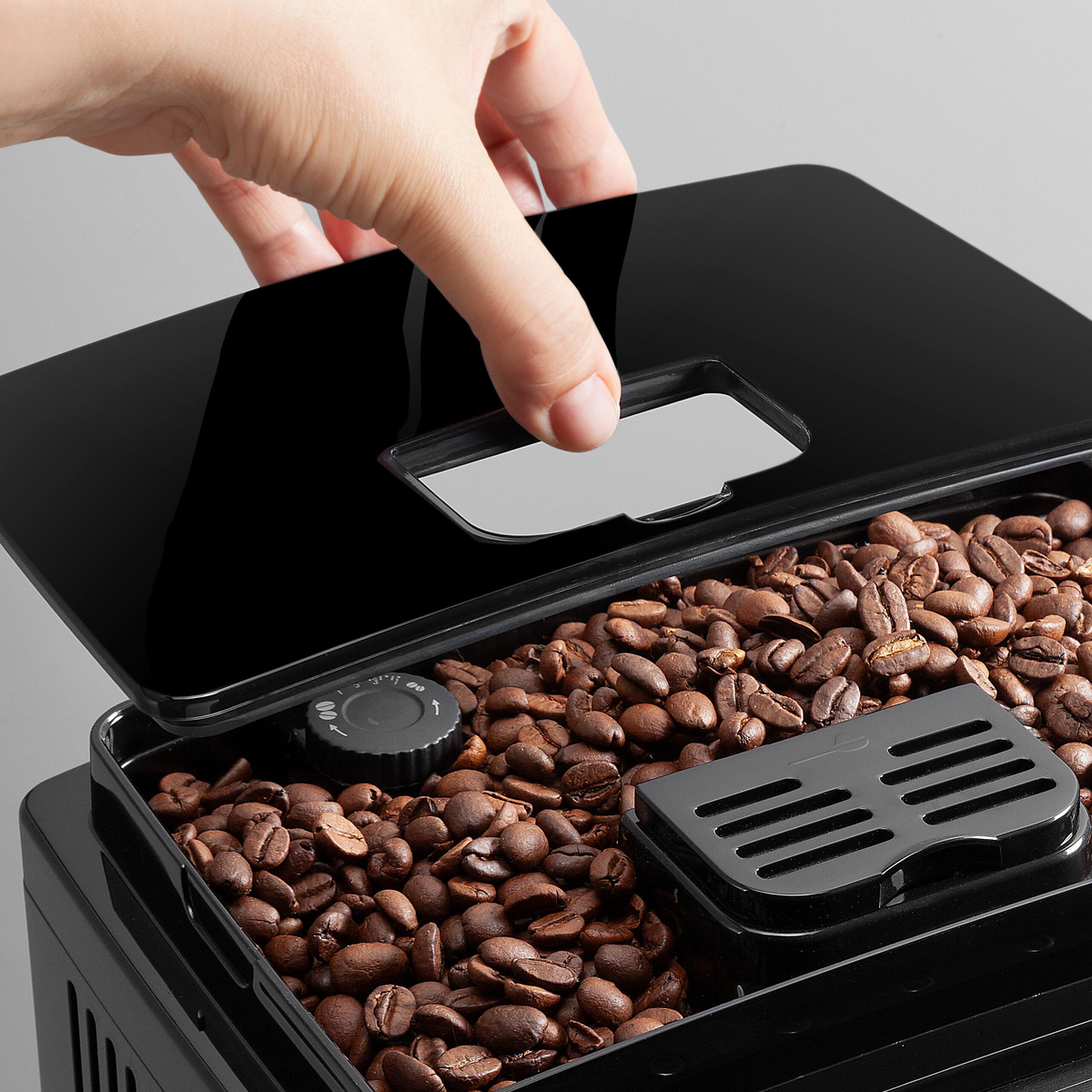 Dłoń otwiera czarną pokrywę ekspresu do kawy. Ziarna wypełniają komorę.