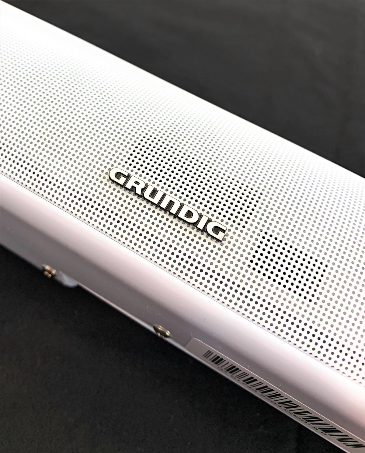 Srebrny soundbar Grundig, z czarnym perforowanym przodem. Słowo Grundig jest na górze.