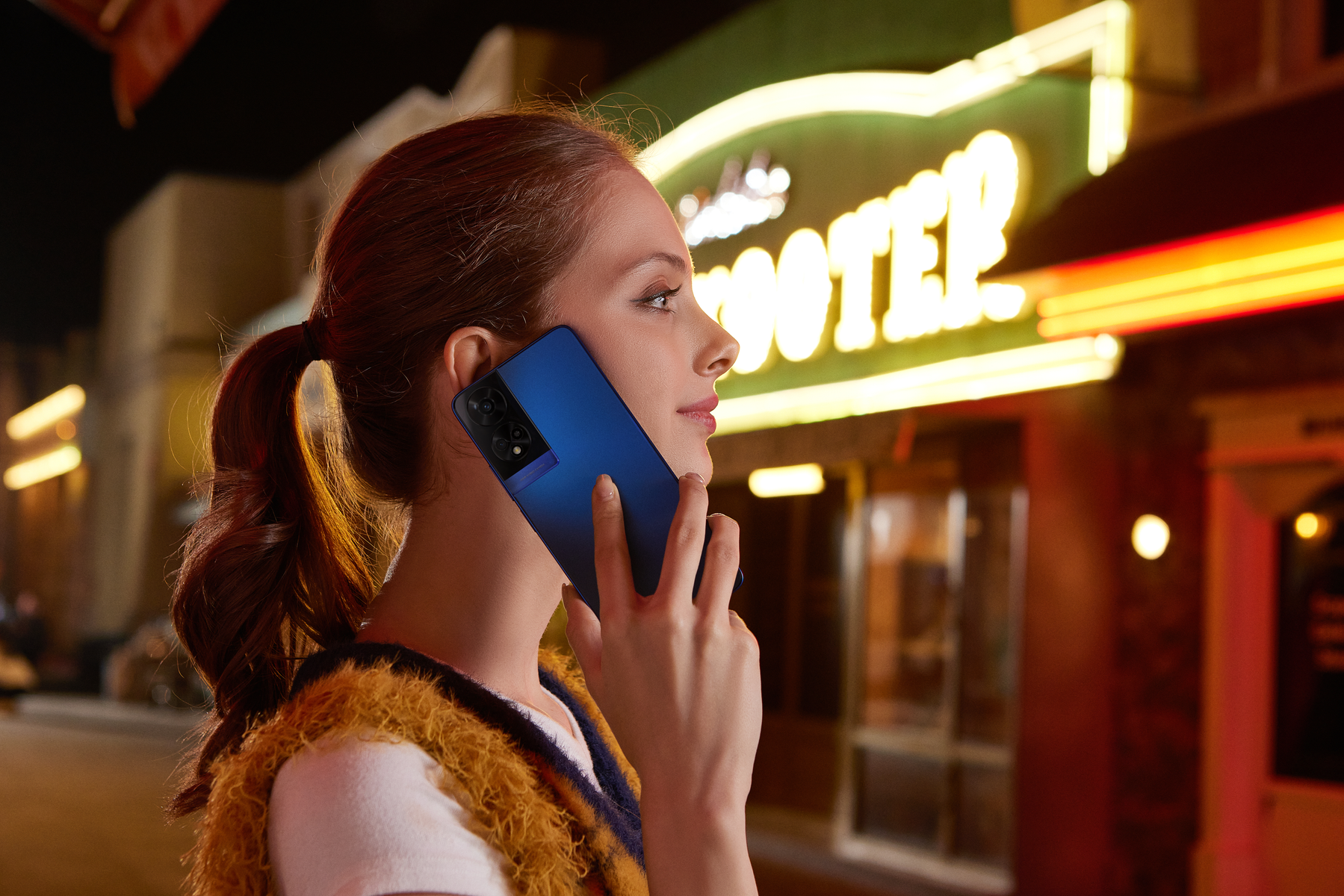 Una mujer sosteniendo un teléfono inteligente azul en su oído, frente a un letrero de 'MOTEL'.