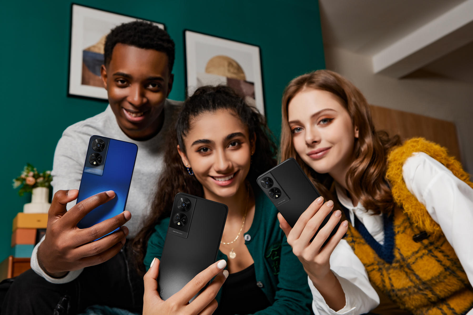 Tres personas sonriendo sostienen teléfonos, uno azul y dos negros, frente a una pared turquesa.