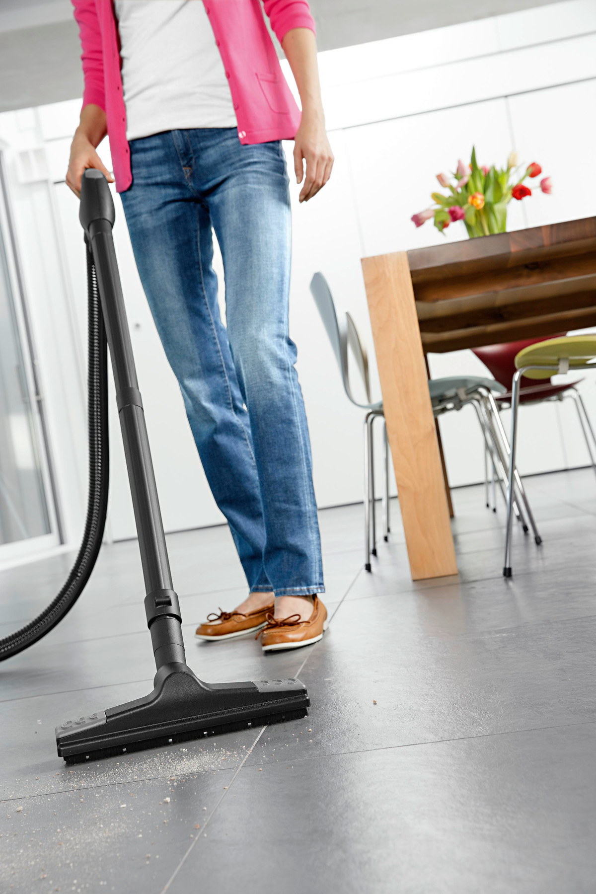 Osoba odkurzająca szarą podłogę z płytek. Kobieta w dżinsach, różowym topie, brązowych butach.