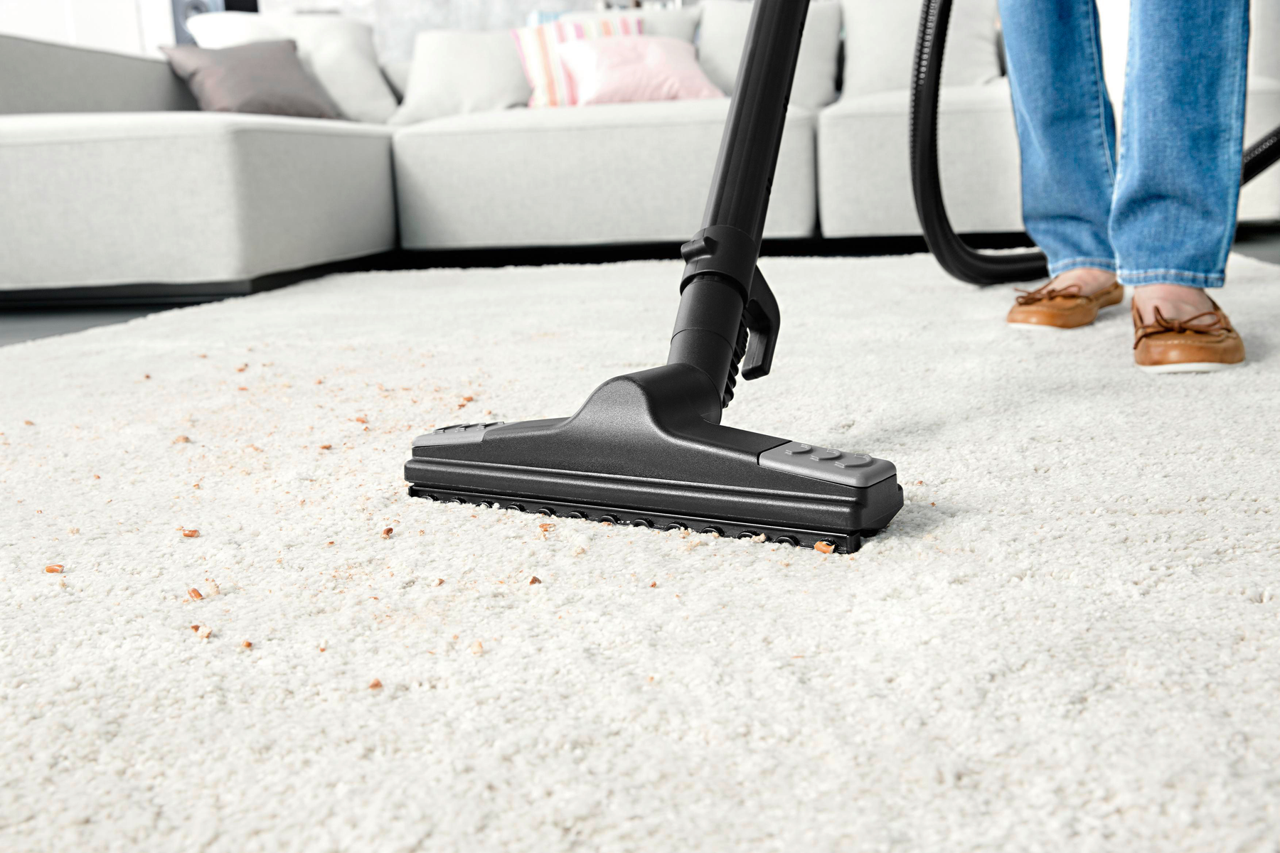 Osoba odkurza biały dywan z resztkami. Głowica odkurzacza znajduje się na dywanie, widoczne są nogi i stopy osoby.