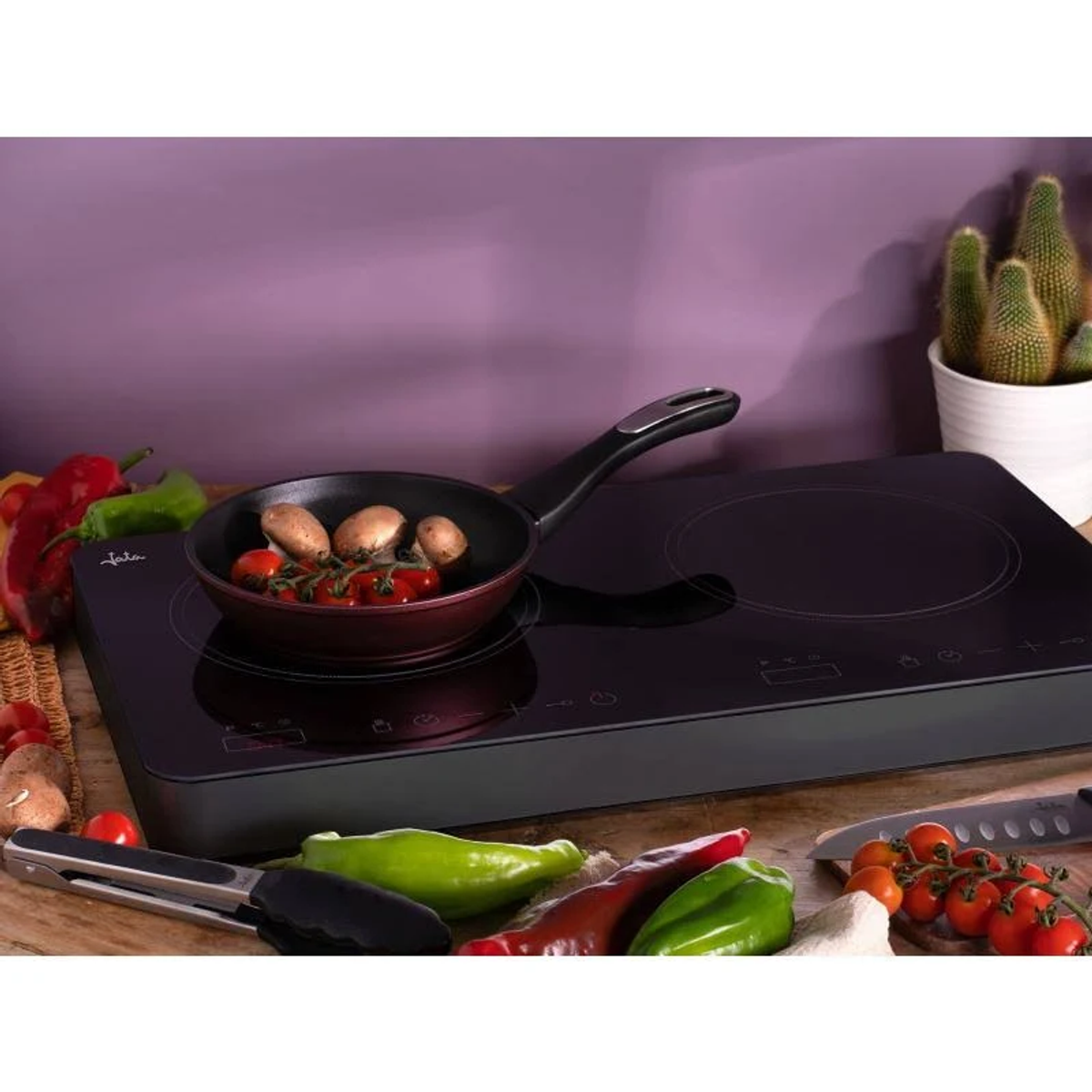 Una placa de inducción negra con una sartén con champiñones y tomates. Verduras y un cuchillo alrededor.