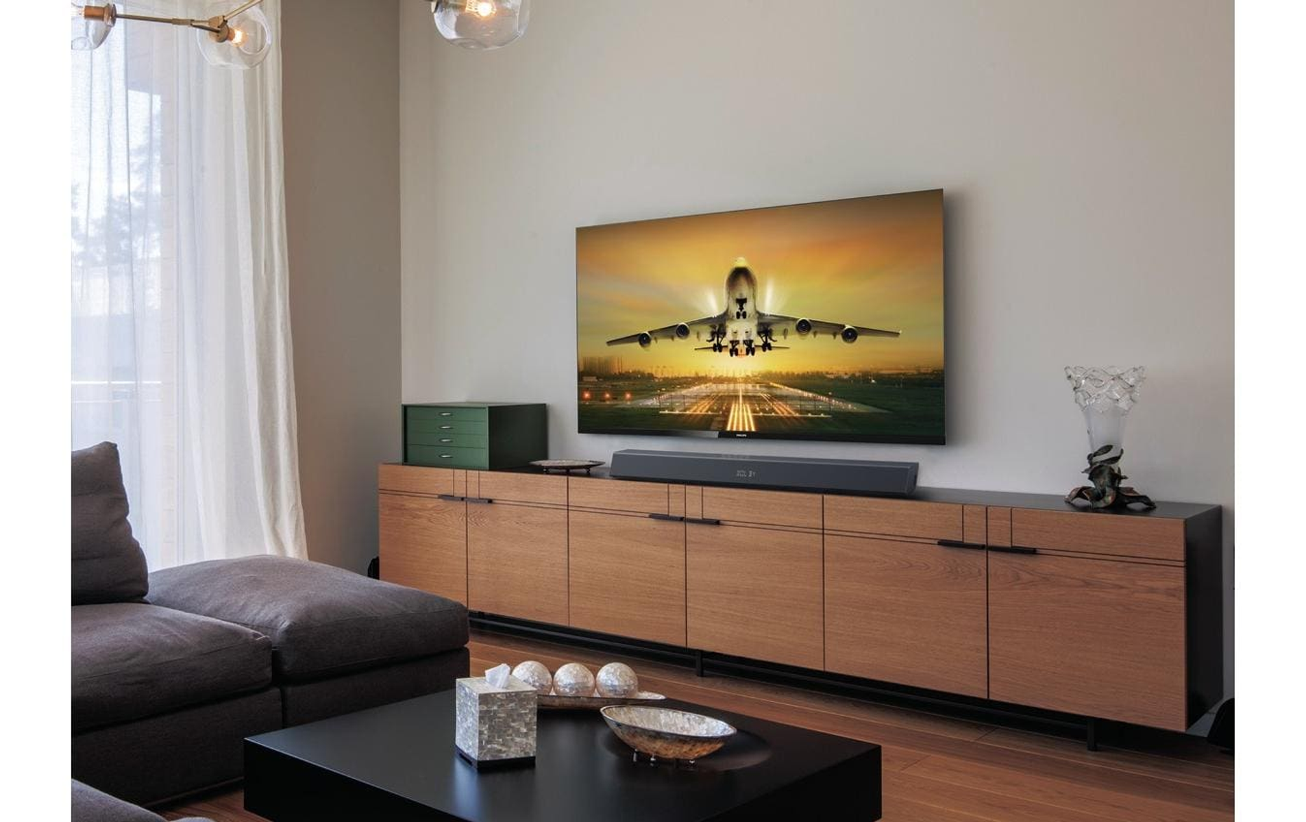 Wohnzimmer mit TV, das eine Flugzeuglandung zeigt. Soundbar unten. Holzschrank, dunkle Möbel.