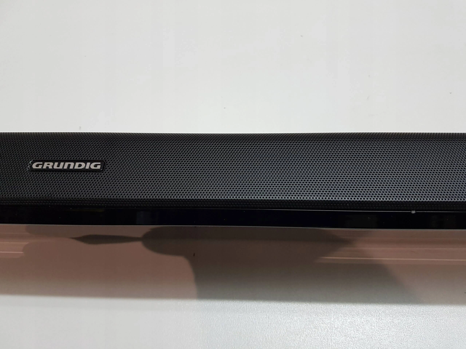 Czarny soundbar z logo Grundig. Soundbar znajduje się na białej powierzchni. Czarne tło.