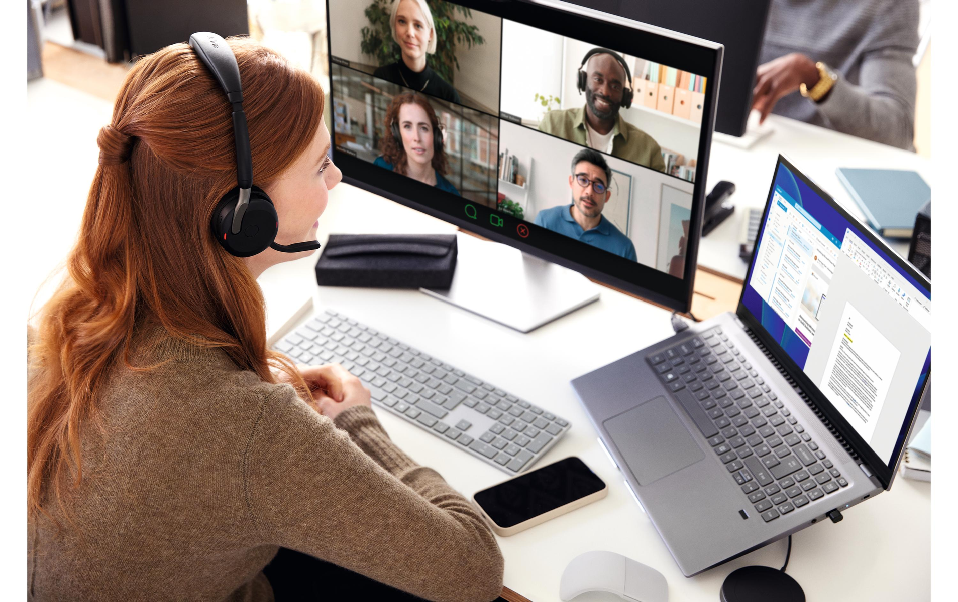 Kobieta w zestawie słuchawkowym podczas rozmowy wideo, przeglądająca ekran z innymi uczestnikami i laptopem.
