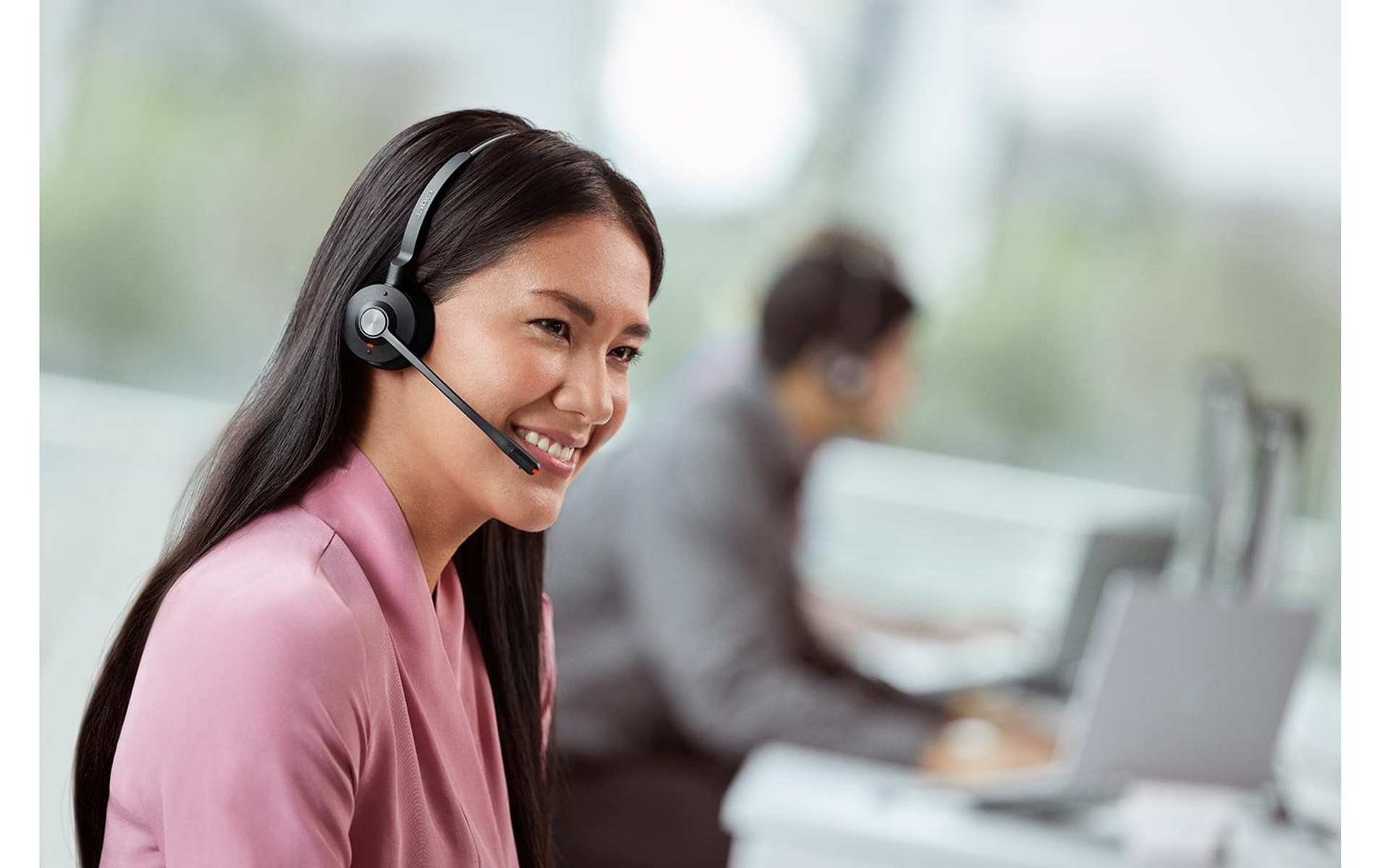 Mujer usando auriculares, sonriendo. Otra persona borrosa en el fondo, trabajando en una laptop.