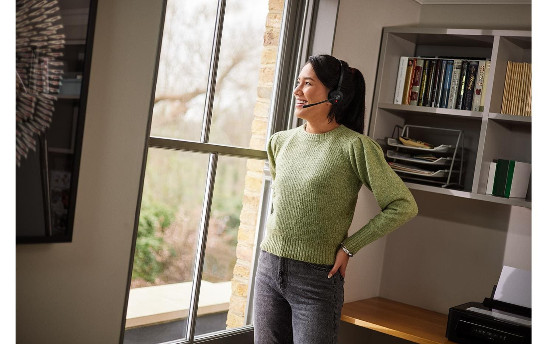 Mujer con auriculares mirando por la ventana, sonriendo, en una oficina en casa.