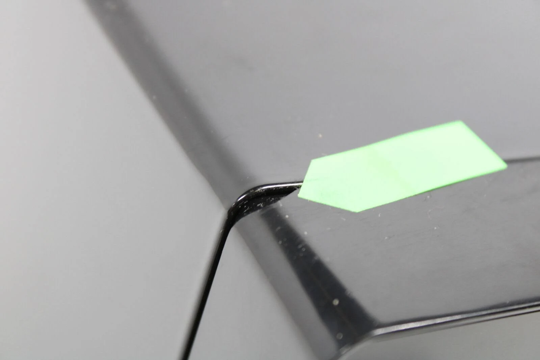 Una esquina de un objeto negro, con una pegatina verde encima. El fondo es gris.