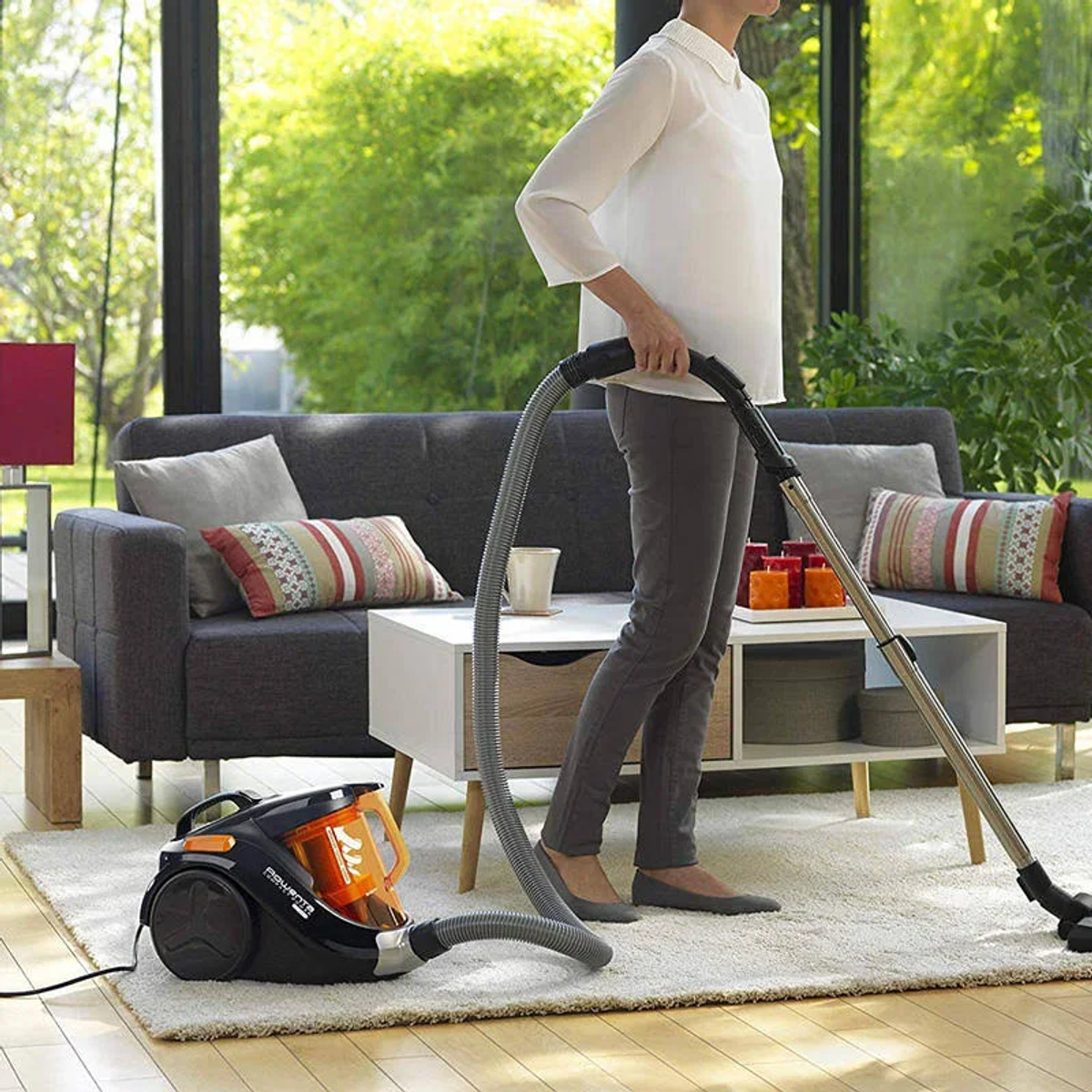 Kobieta odkurzająca dywan w salonie z sofą, stolikiem kawowym i roślinami.