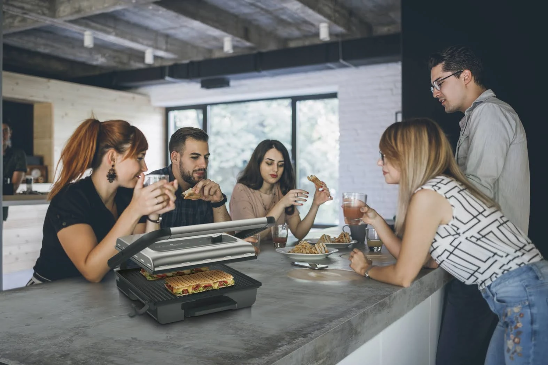 Gruppo di persone intorno a un tavolo con un'affettatrice, mangiano panini.