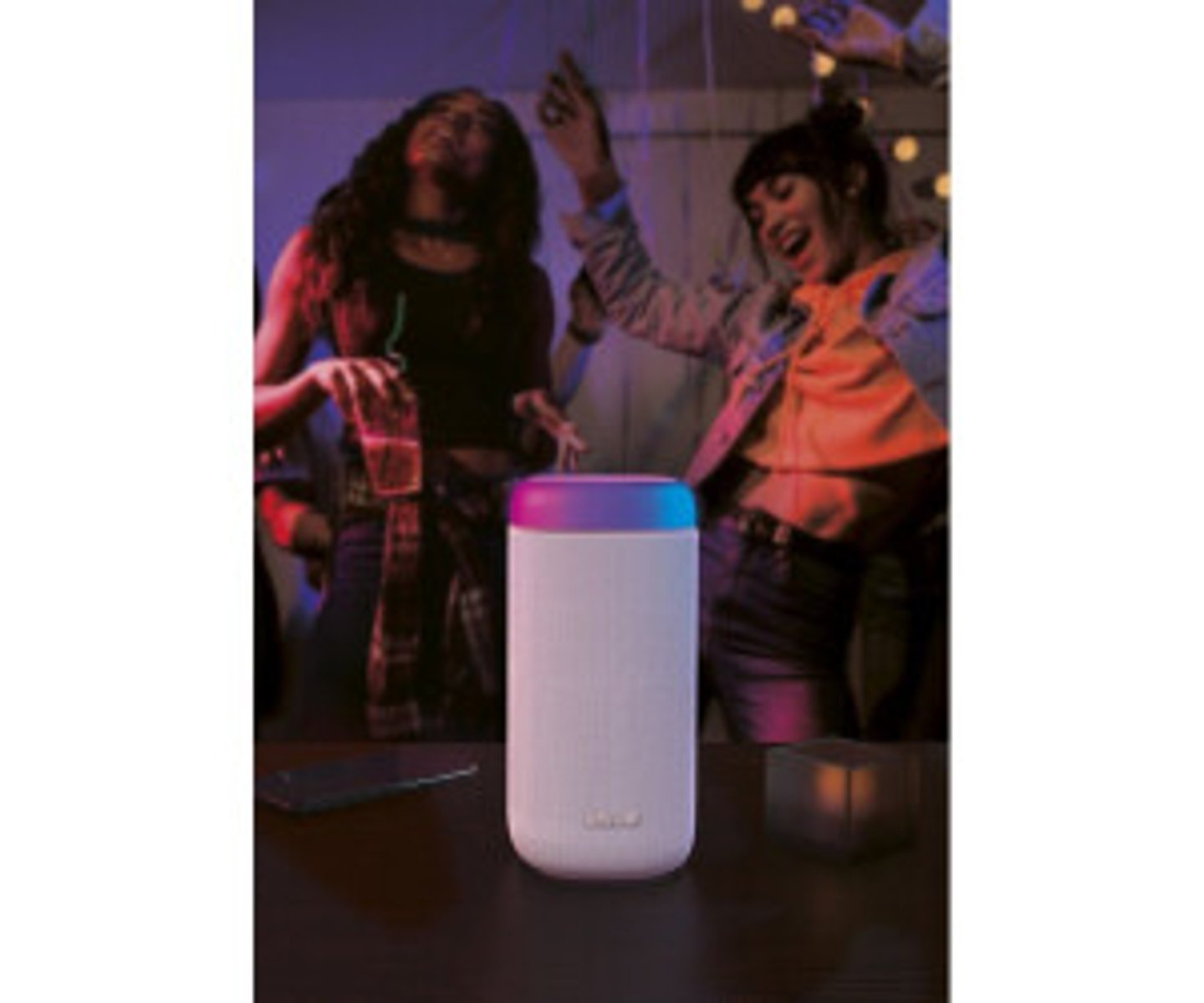 Biały głośnik z kolorowymi światłami, dwie osoby tańczące w tle.