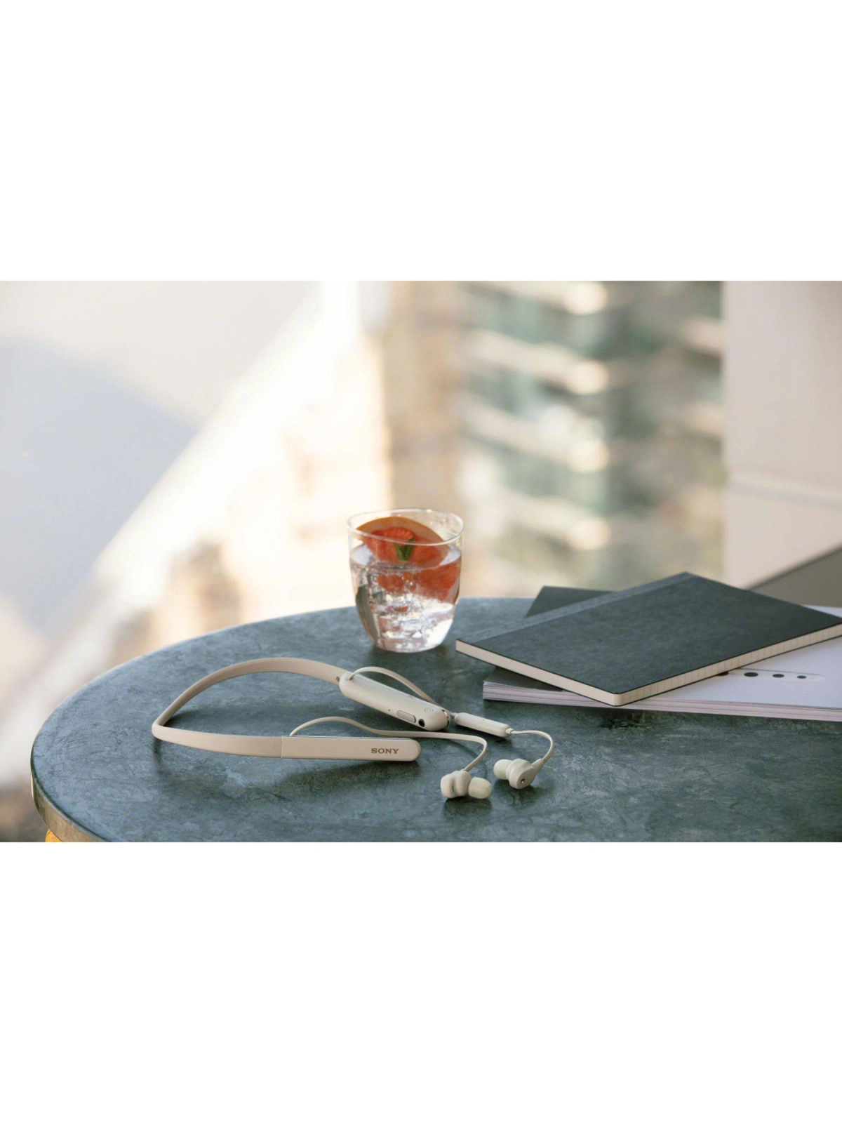 Headphones, water glass, and notebooks are on a table. Background: city view.