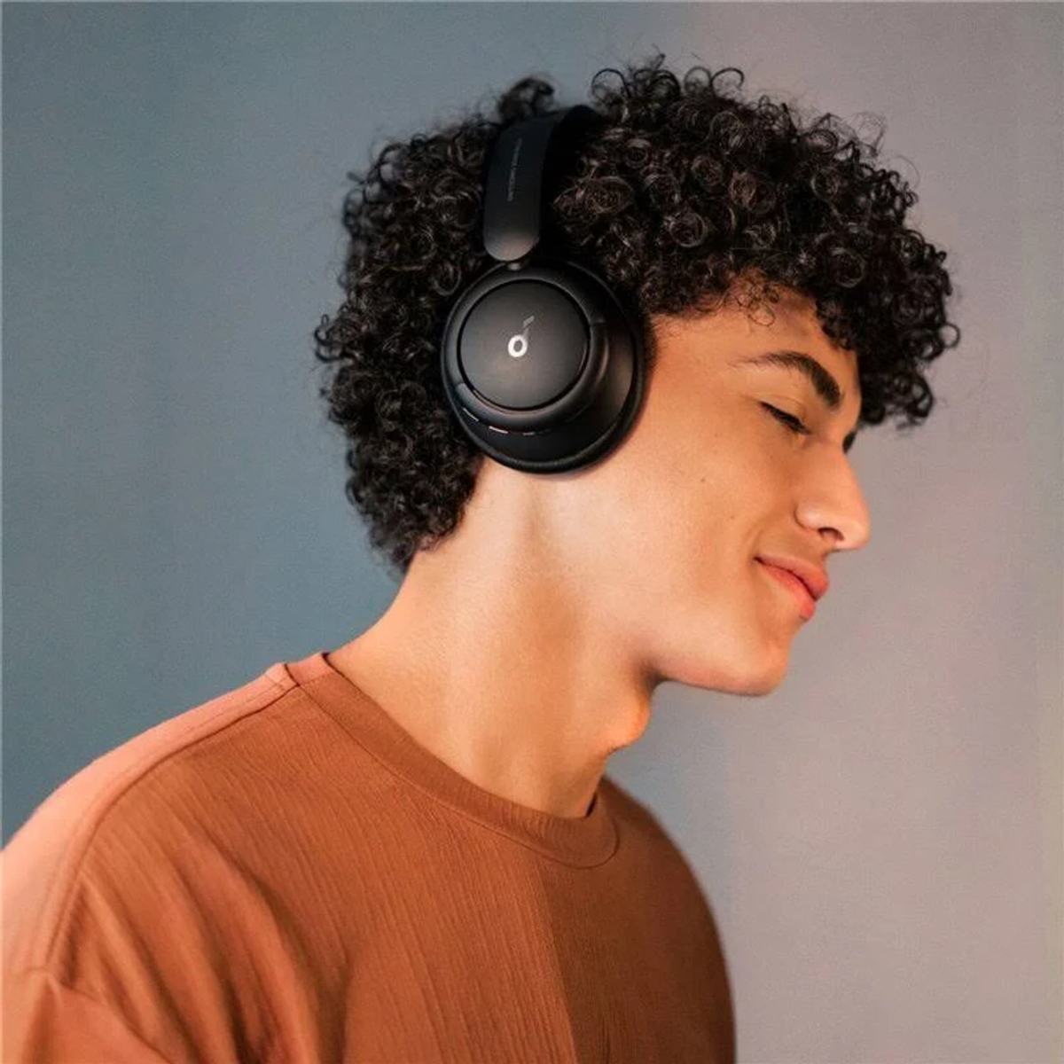 Una persona con auriculares negros, sonriendo, con una camisa marrón. Fondo gris.