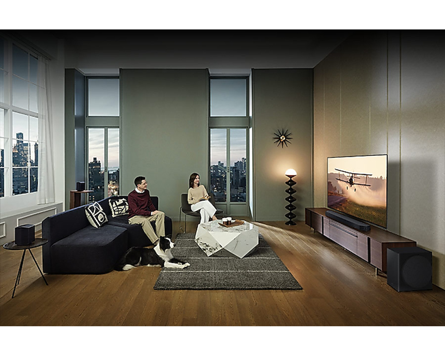 Ein Wohnzimmer mit einem Paar, einem Hund und einem Fernseher mit Landschaft.