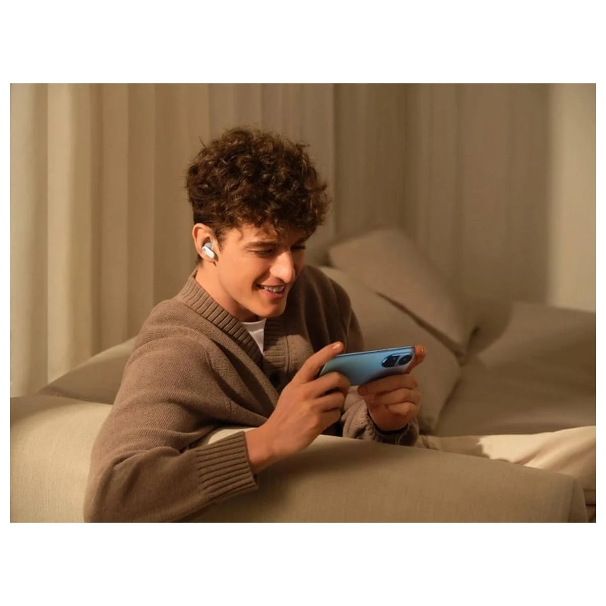 Un joven sonríe mientras juega en un teléfono. Lleva auriculares y está sentado en un sofá.