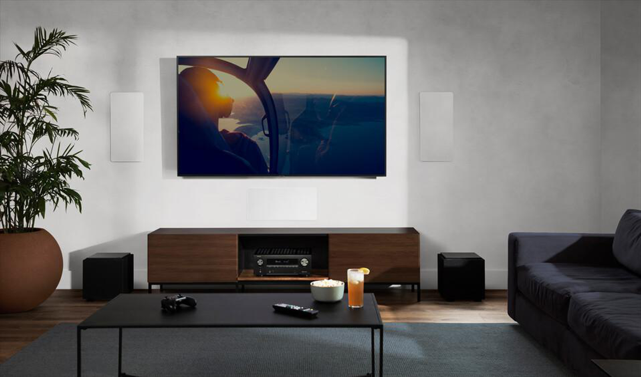 Wohnzimmer mit TV-Gerät, das eine Hubschrauberszene zeigt, Lautsprechern, Sofa und Couchtisch mit Getränken.