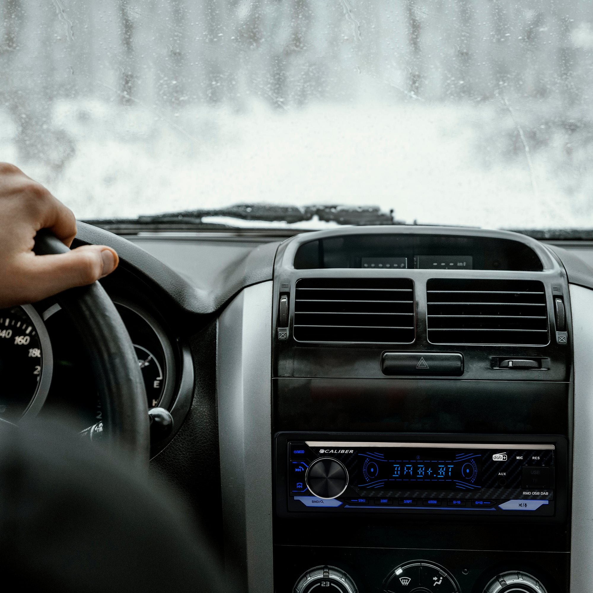 Autointerieur mit einer Hand am Lenkrad. Ein Radio ist sichtbar. Die Szene ist verschwommen, was Bewegung und Wetter andeutet.