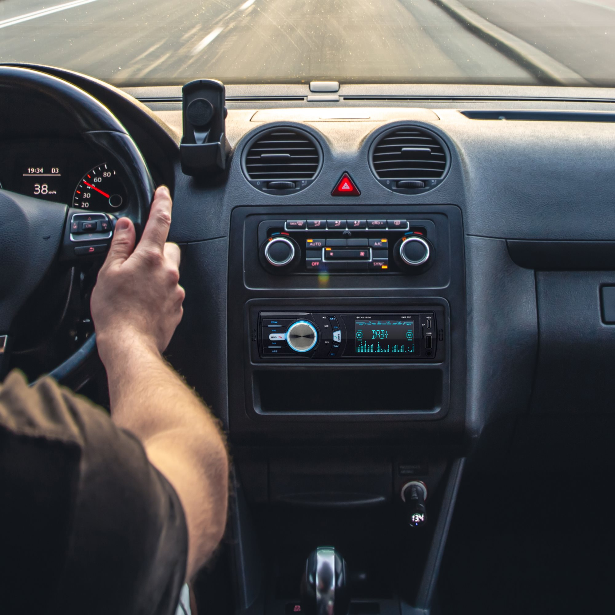 Handen van de bestuurder op het stuur van een auto. Het dashboard heeft een stereo en een display.